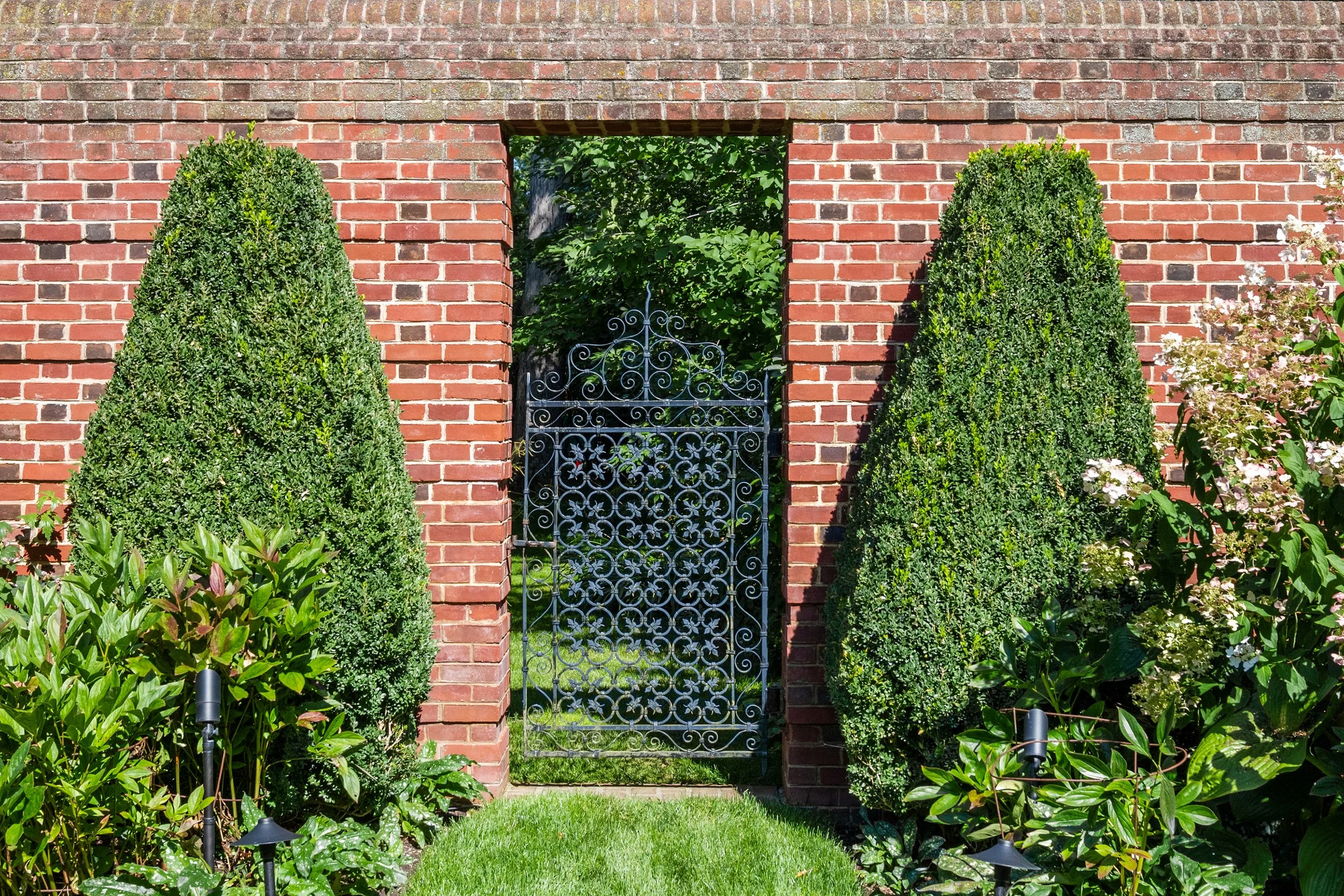 A brick wall with a rectangular opening in the middle, featuring an ornate iron gate. There are two tall, cone-shaped green bushes on either side of the opening, and flowering plants with pink and white blooms on the right, along with lush green gras