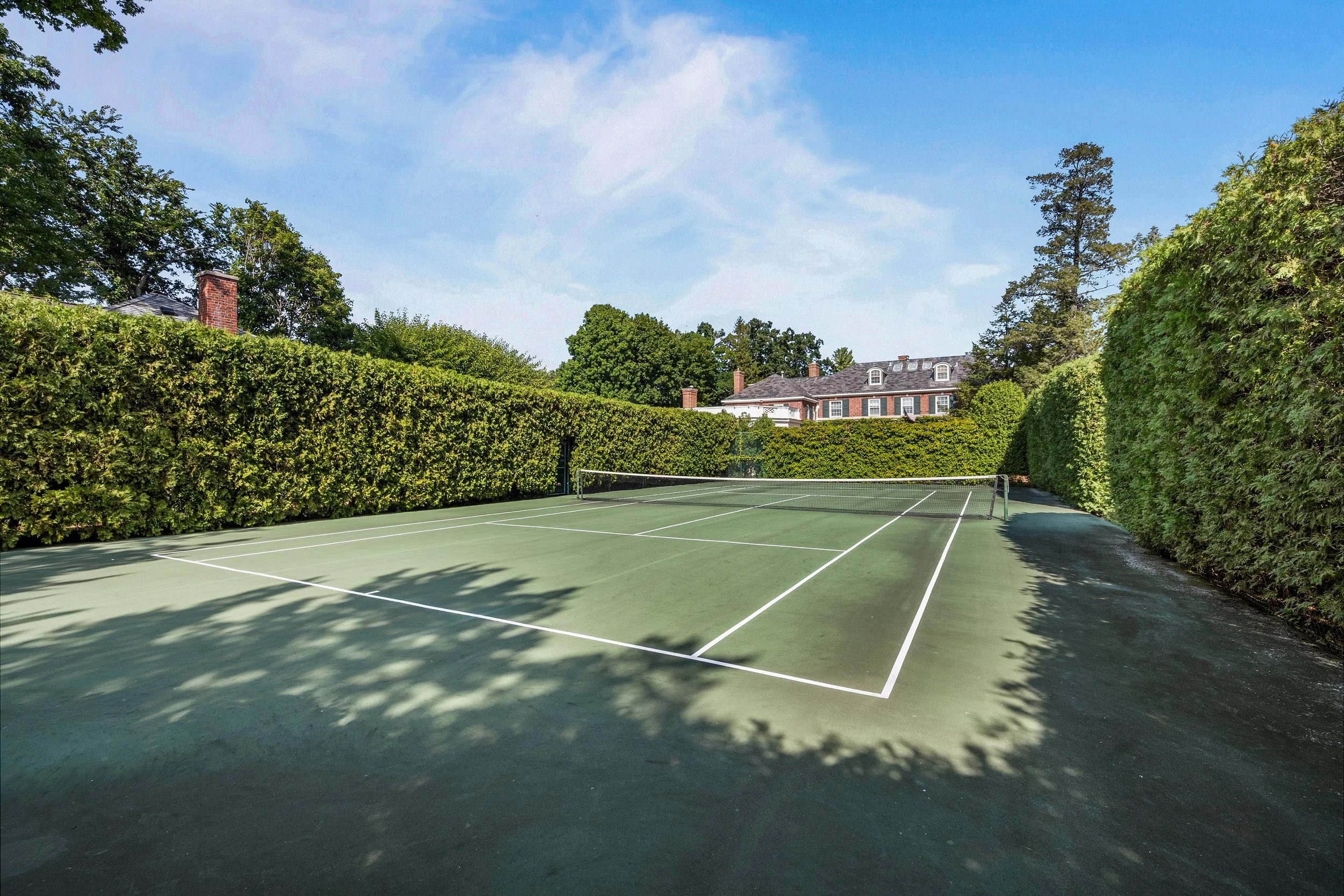 An empty outdoor tennis court surrounded by tall green hedges under a blue sky with some clouds.