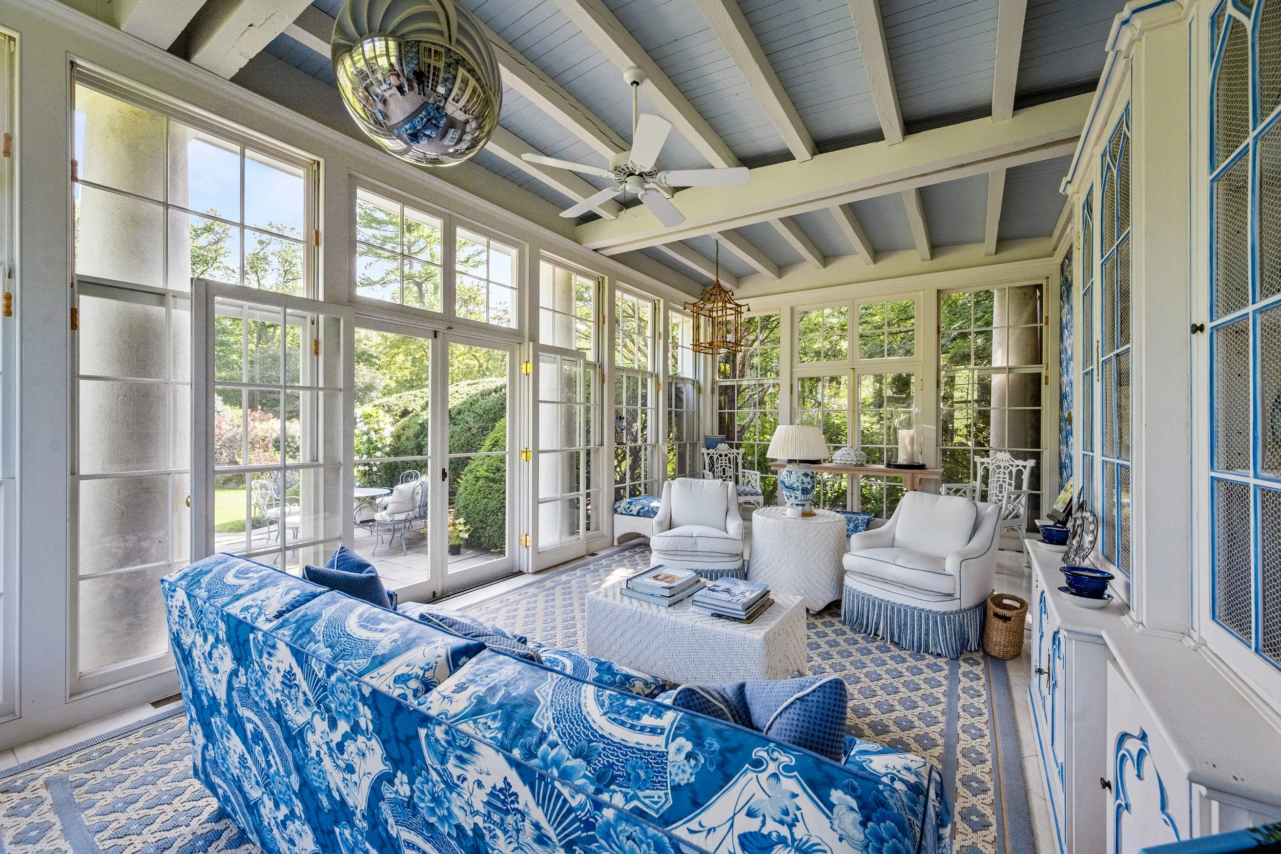 Bright sunroom with large glass windows, white and blue furniture, patterned blue and white curtains, and a view of a lush garden outside.