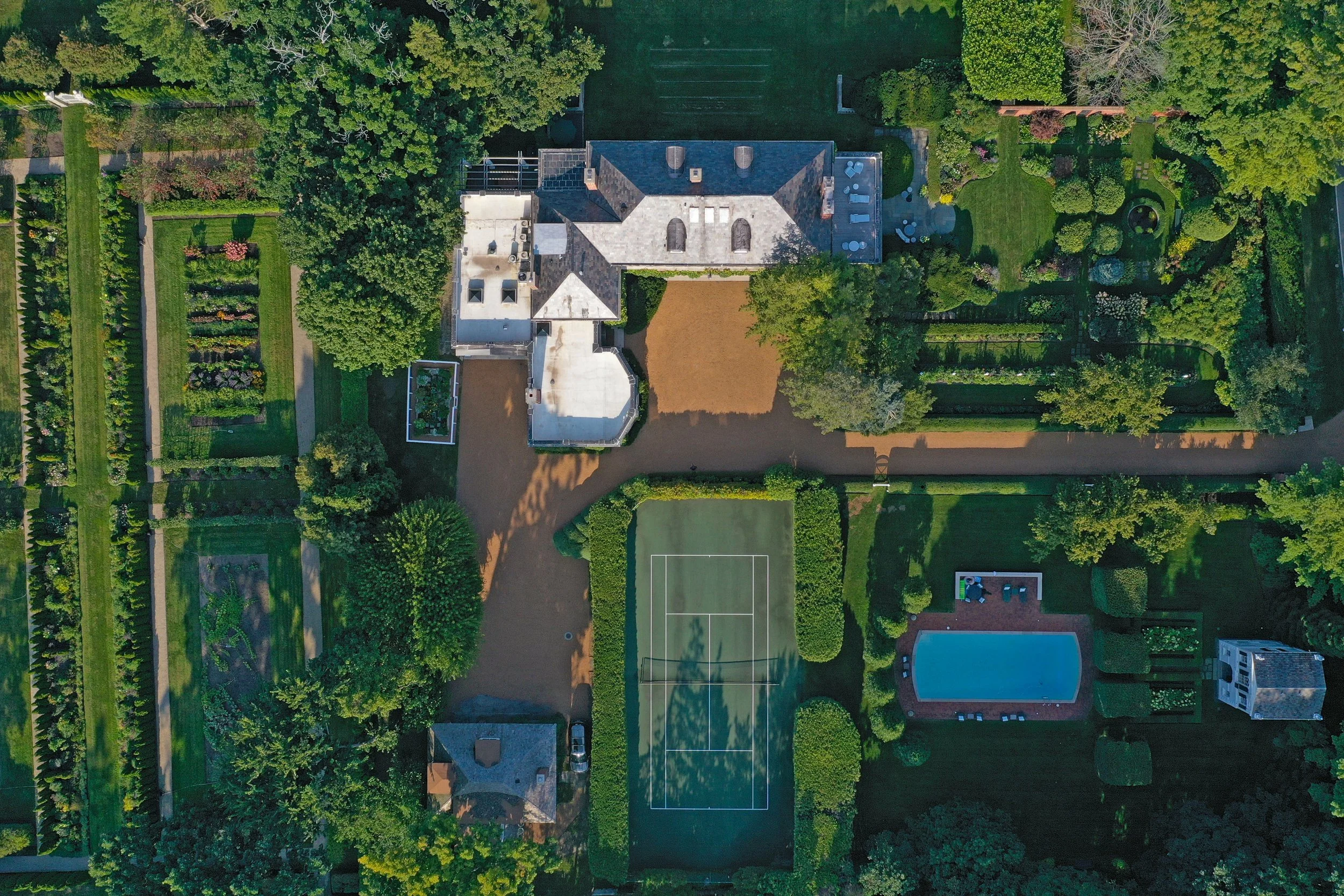 An aerial view of a large house with a gray roof surrounded by lush green trees and a well-maintained garden. The property includes a tennis court, a swimming pool, and multiple outdoor seating areas.