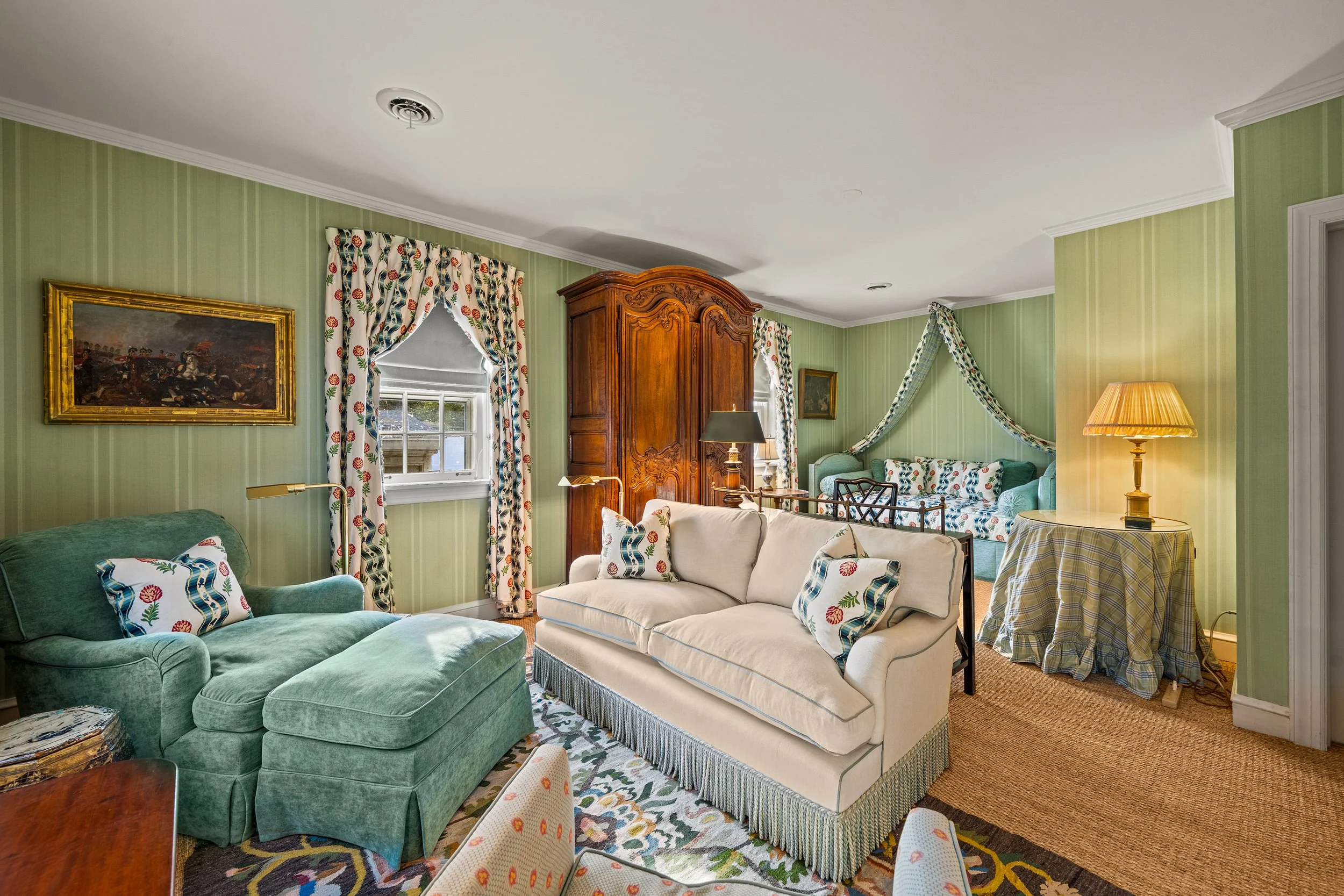 Living room with green striped wallpaper, beige sofa, green armchair, wooden armoire, floral curtains, table lamps, and decorative pillows