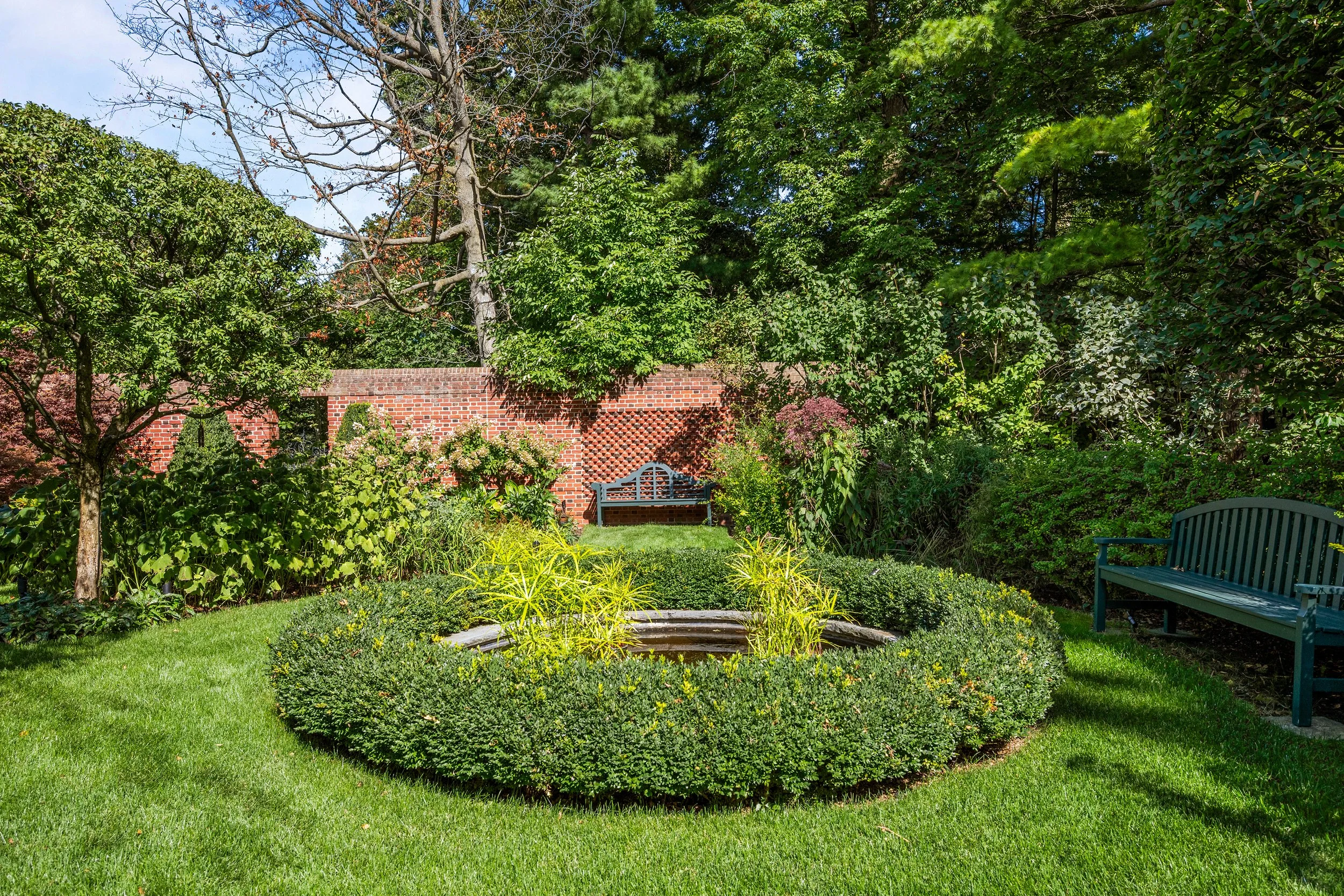 A lush garden with green grass, trees, and bushes surrounds a small circular pond with yellow plants. There are two benches, one in the background near a red brick wall and another on the right side.