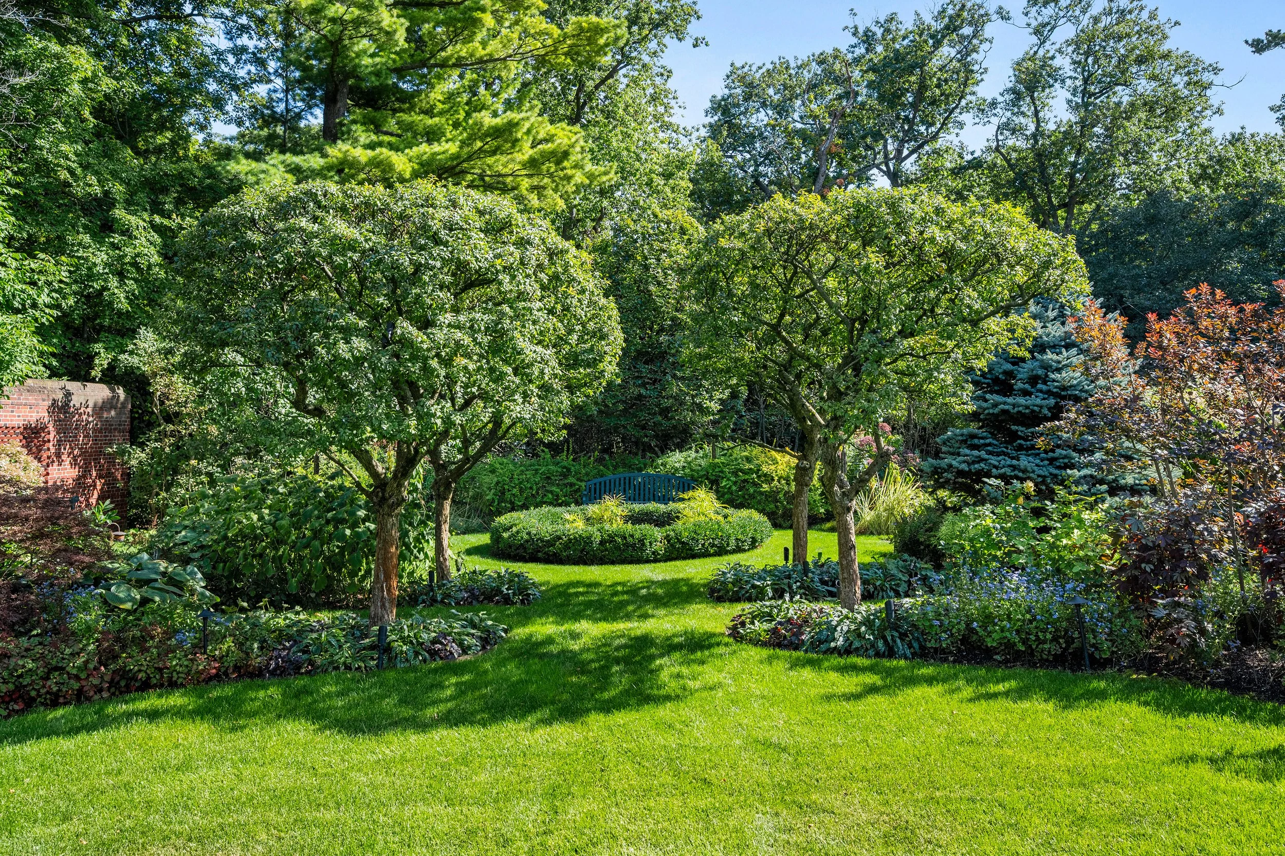 A lush, well-maintained garden with green grass, two trimmed trees, and a variety of shrubs and plants. There is a blue bench among the foliage and a brick wall partially visible on the left side.