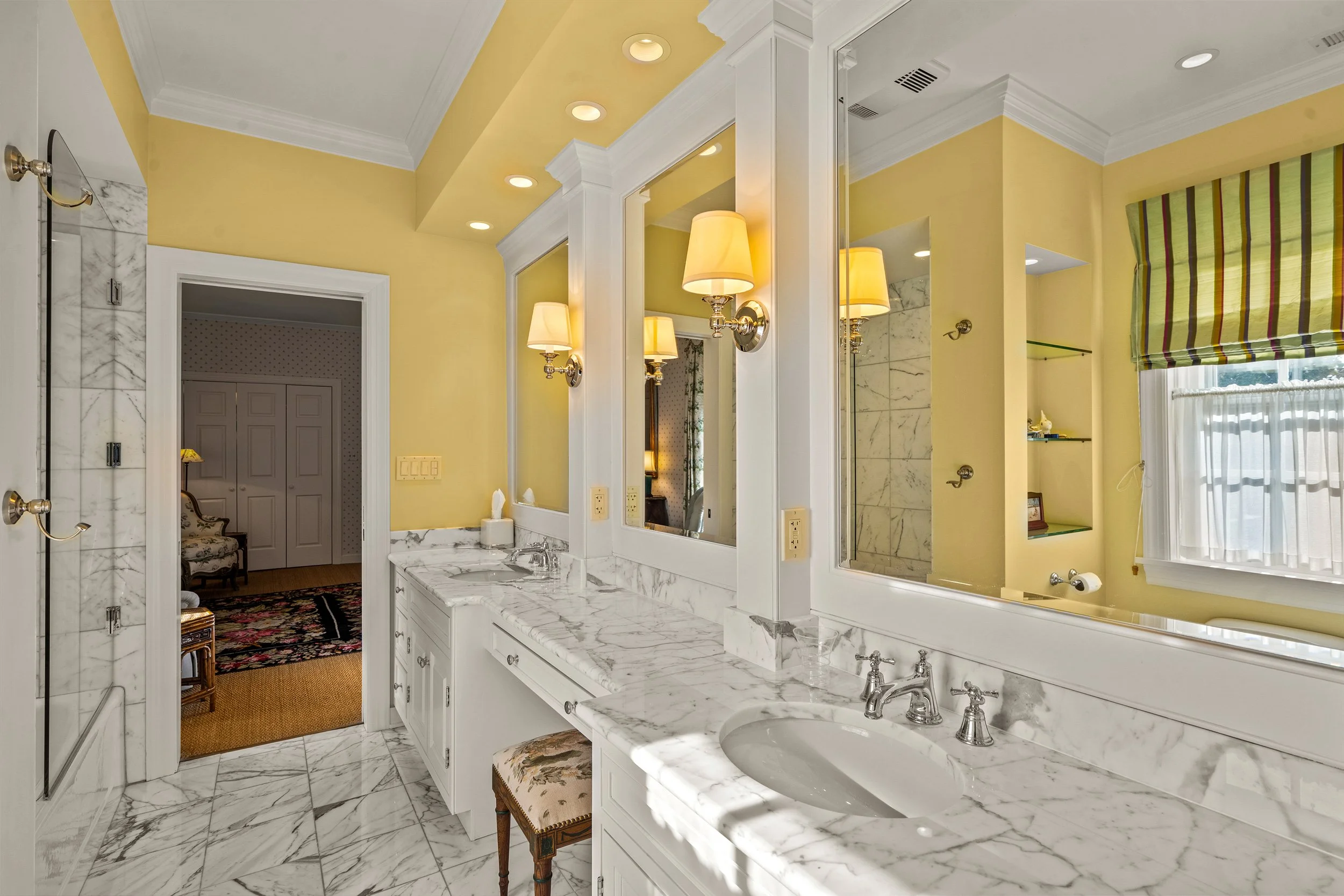 Bathroom with yellow walls, marble countertop, and large mirrors. Double sinks, wall-mounted lamps, and a window with a striped valance. An adjacent room with floral armchair and closet visible.