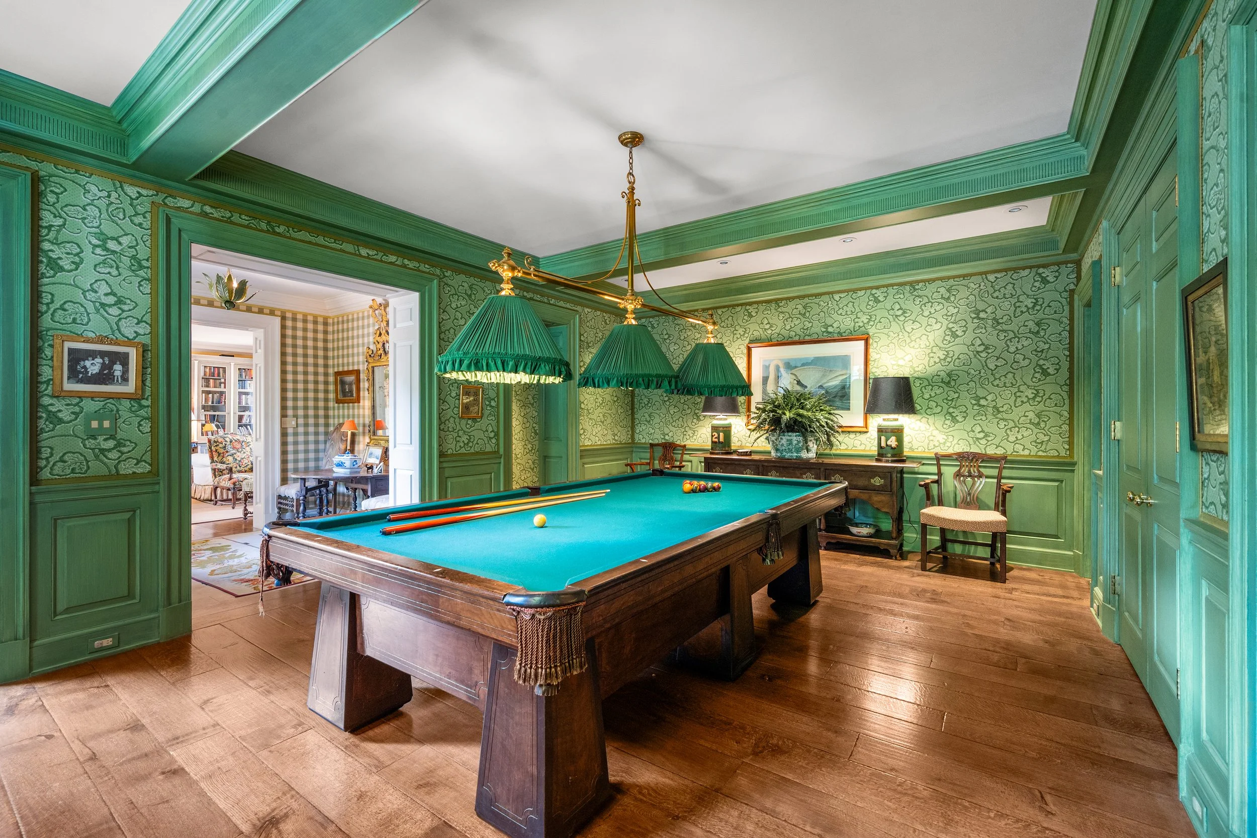 A room with green patterned wallpaper, a wooden pool table with green felt, pool balls and cues, and a chandelier with green lampshades.