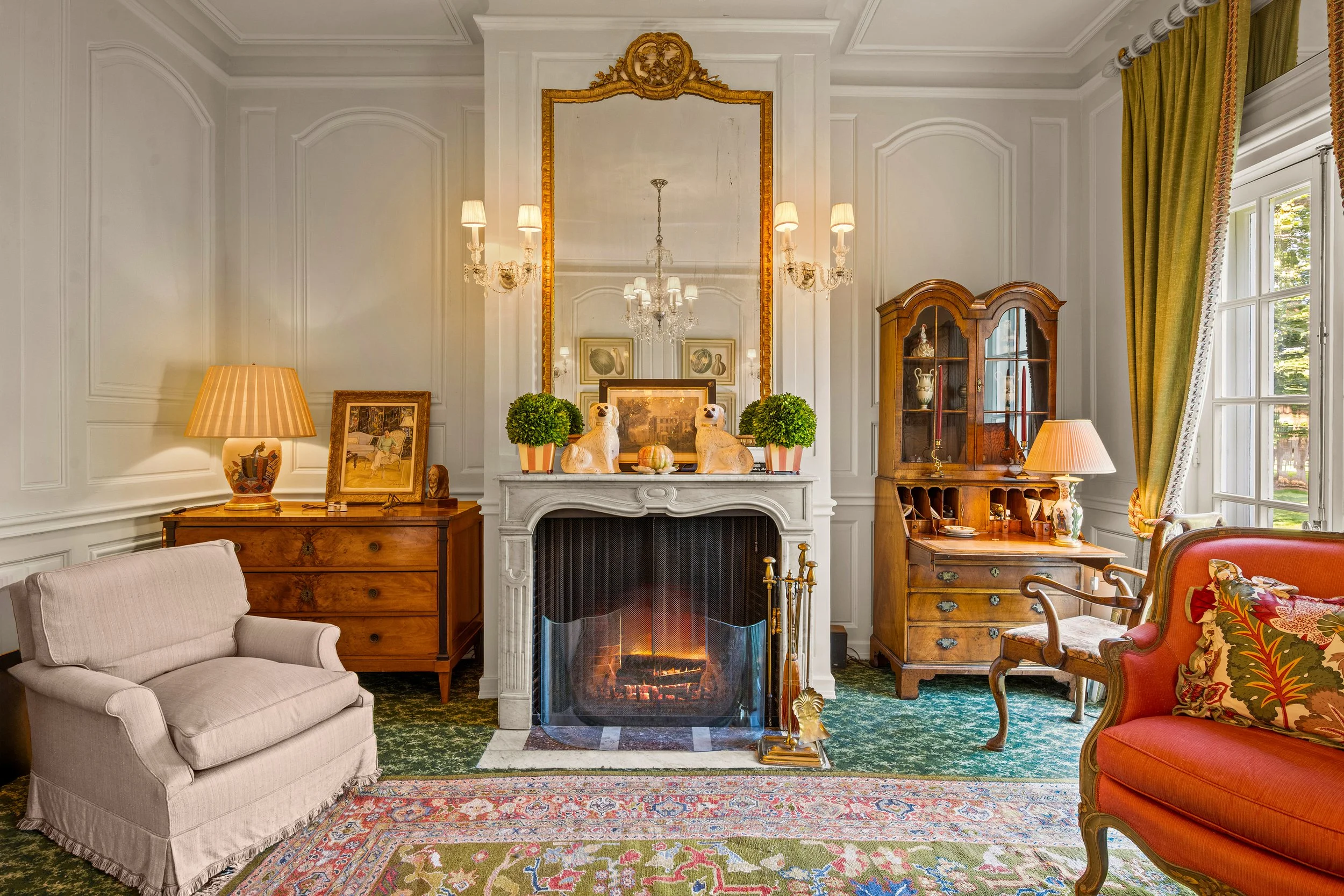 Cozy living room with white fireplace, decorated with two dog figurines, framed pictures, and small green plants. To the left, a beige armchair and wooden dresser with a lamp and framed artwork. To the right, a wooden display cabinet, a side table wi