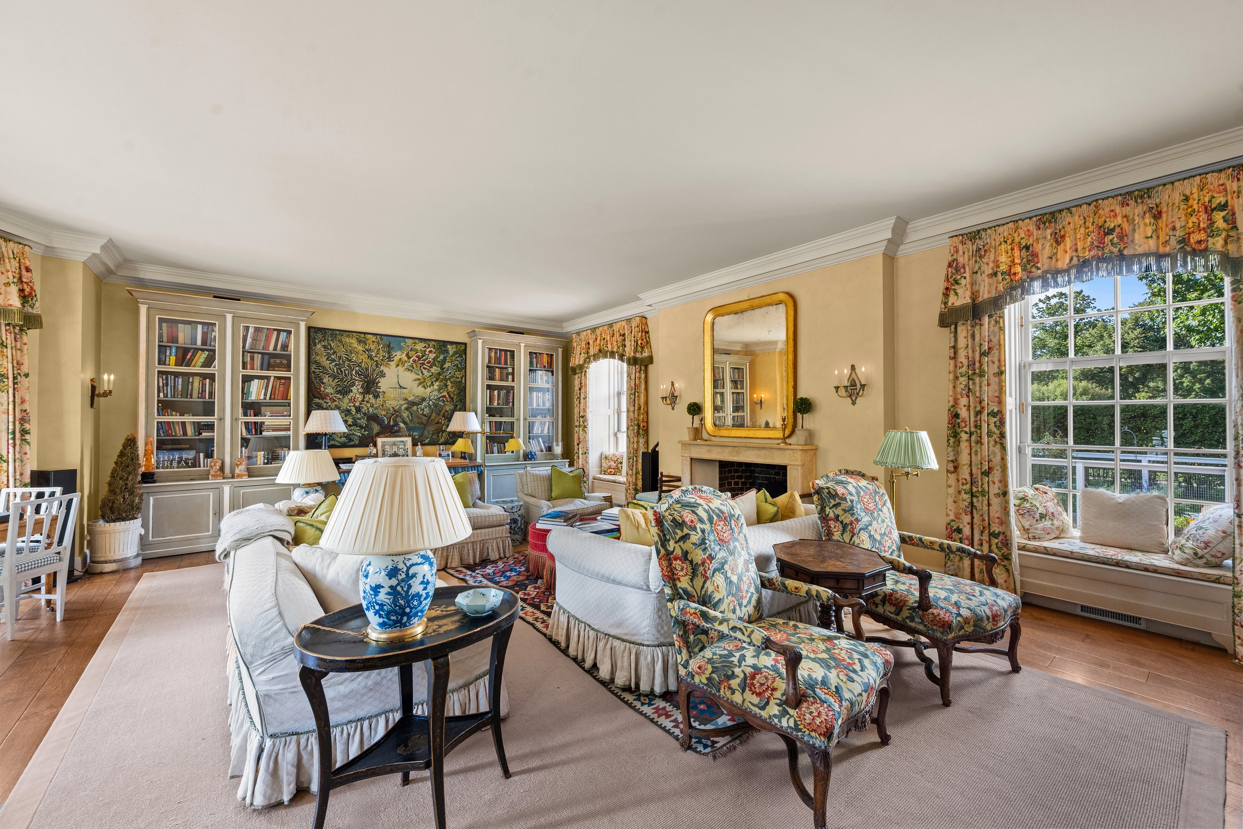 A spacious, well-lit living room with large windows, floral curtains, a white couch, floral armchairs, a fireplace with a large gold-framed mirror, and built-in bookshelves filled with books and decorative items.