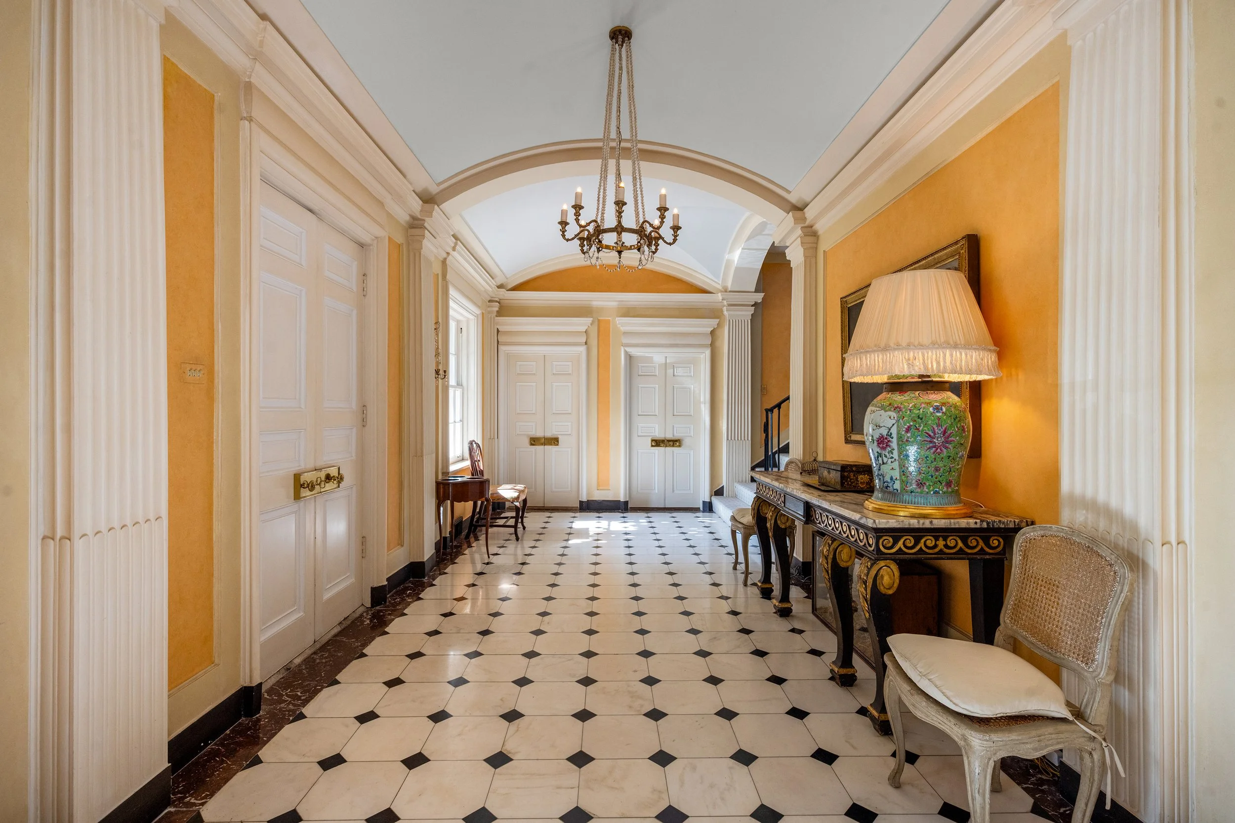 Elegant hallway with yellow walls, white paneled doors, black and white patterned tile floor, chandelier, large floral table lamp, and vintage chairs.