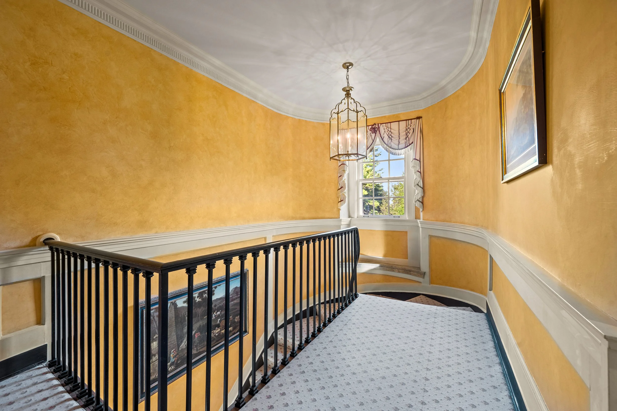 Yellow walls with white crown molding, a window with patterned curtains, a chandelier, and framed artwork in a residential hallway.