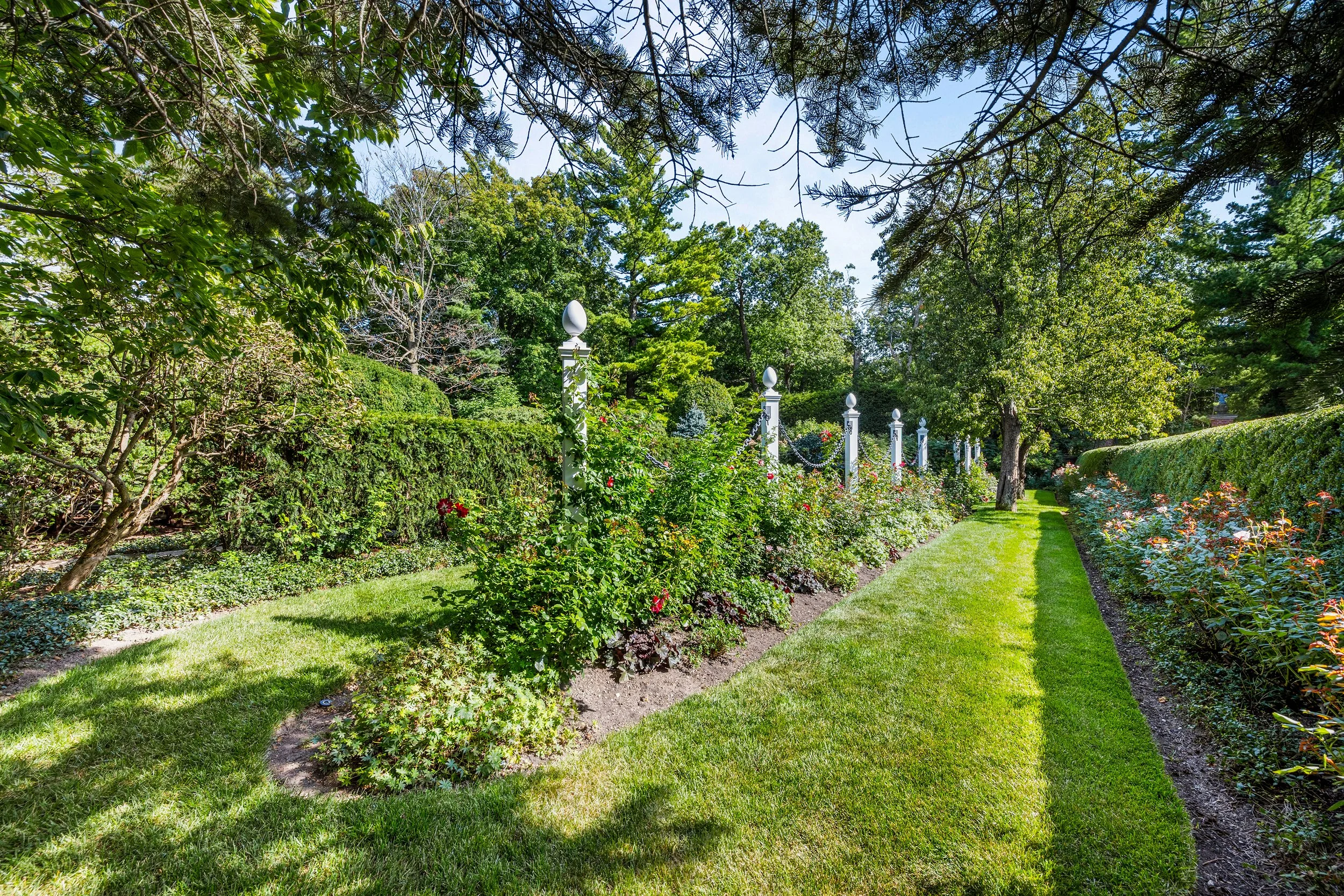 A lush garden with a neatly mowed lawn, rose bushes, and tall trees under a blue sky.