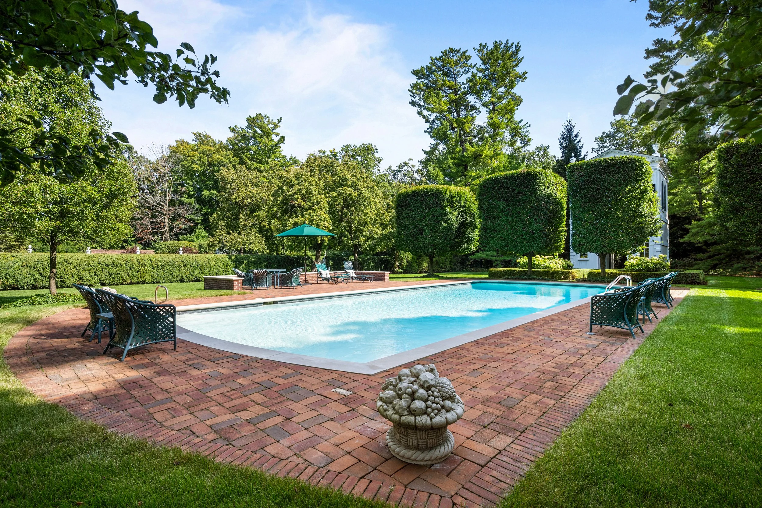 A backyard with a swimming pool surrounded by green grass, trees, and shrubbery, with benches and a table with an umbrella.