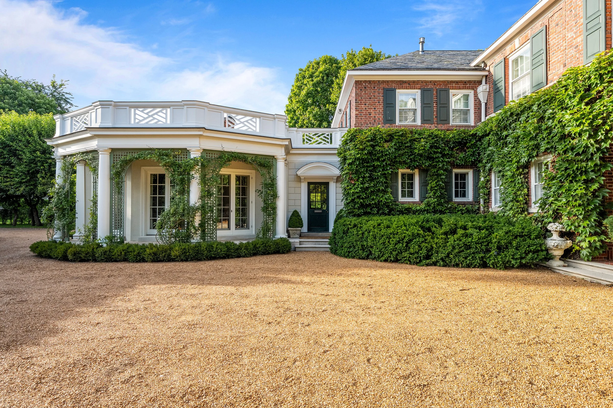 Front view of a large house with a brick and white exterior, ivy-covered walls, multiple windows, a gravel driveway, and a clear blue sky.