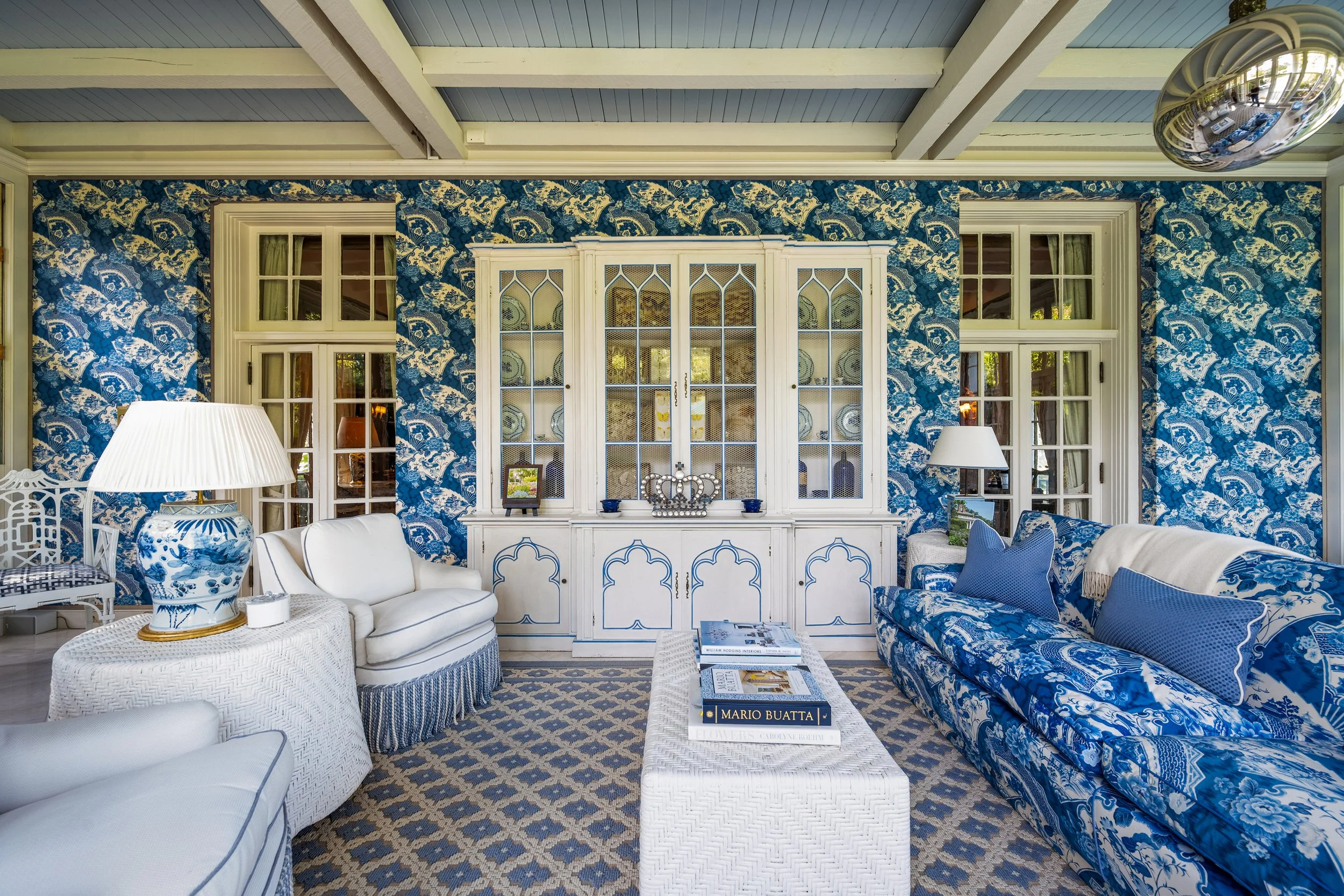 Living room with patterned blue wallpaper, white cabinets, and sofas with blue and white upholstery, large windows, and a patterned carpet.