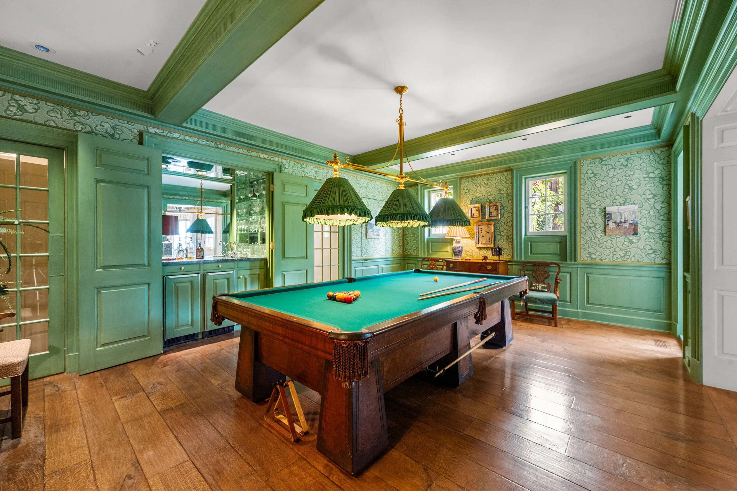 A vintage green-walled game room with a billiards table, brass hanging lamps with green shades, and hardwood flooring