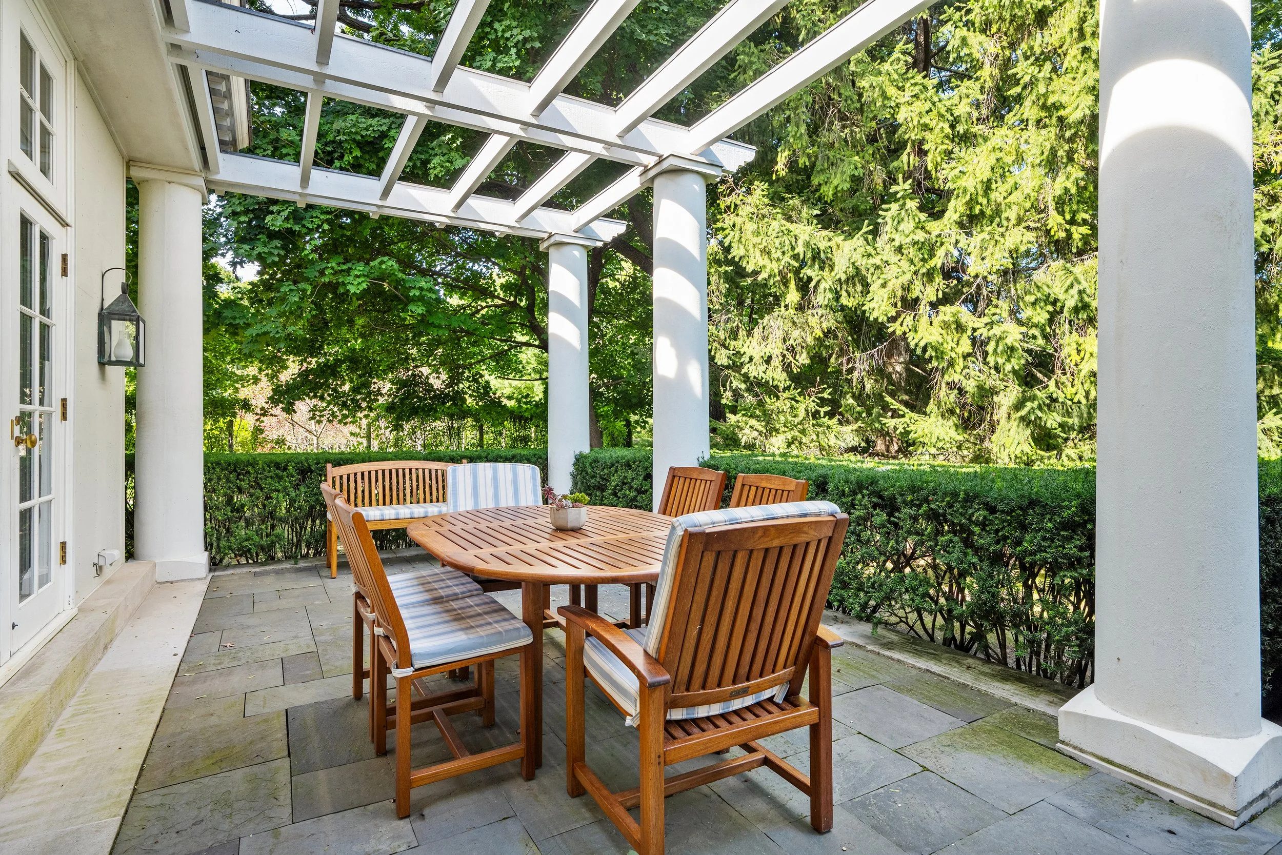Outdoor patio with wooden table and chairs under a white pergola, surrounded by green shrubbery and trees.