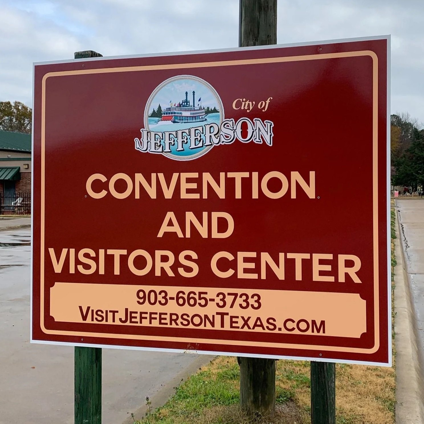 Sign for Jefferson city’s convention and visitors center with phone number and website, near sidewalk with trees and street in background.