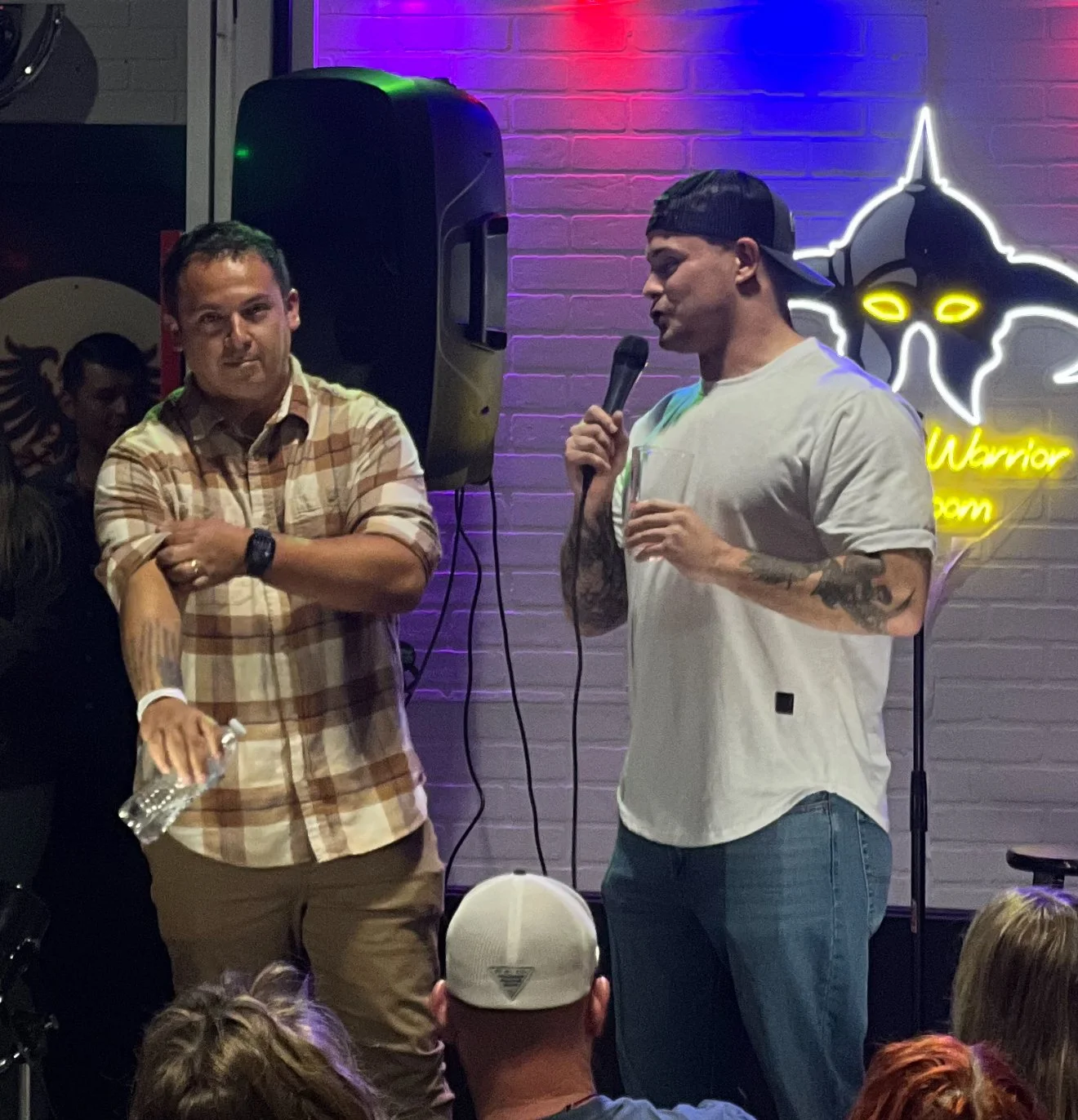 Two men are on stage during a comedy or speaking event; one holds a microphone and a glass, the other stands with crossed arms, in front of a neon sign that says 'Warrior' with a stylized bat or animal logo behind them.
