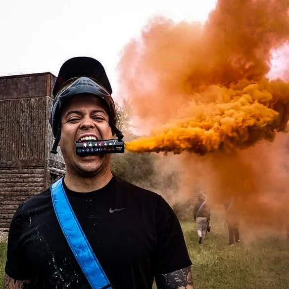A man in a black shirt and helmet is smiling while holding a smoke emitter in his mouth, emitting orange smoke outdoors with other people in the background.