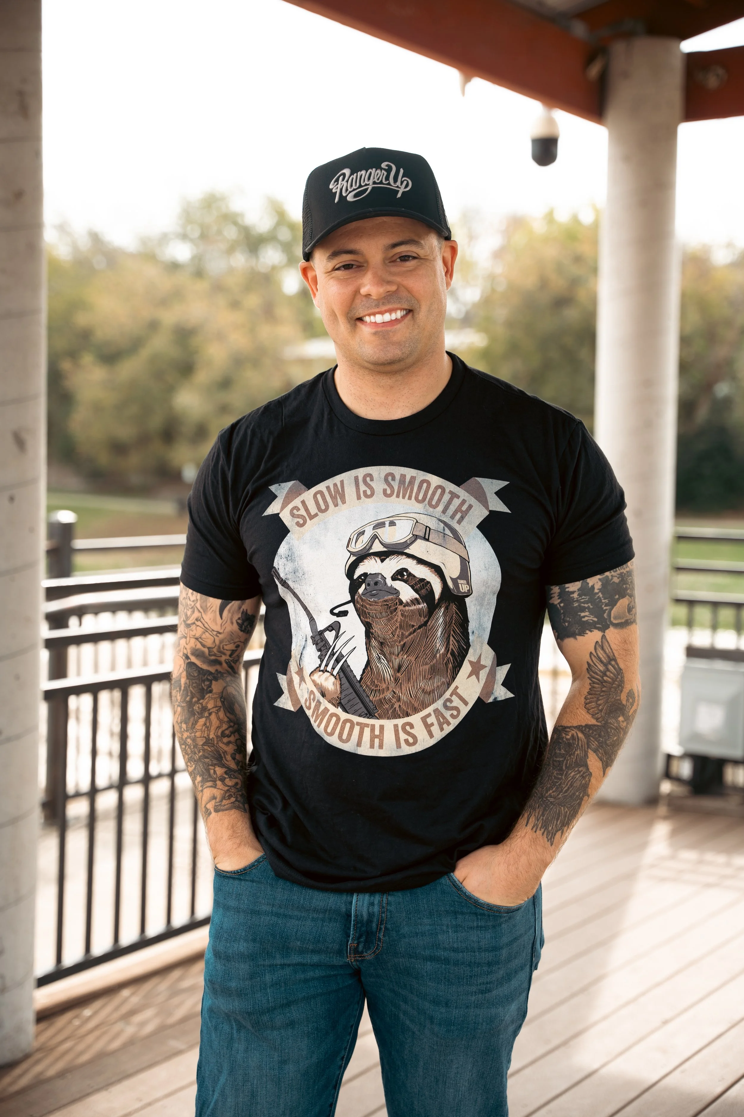 A smiling man with tattoos wearing a black baseball cap and a black t-shirt with a sloth graphic, standing outdoors on a wooden deck with trees in the background.