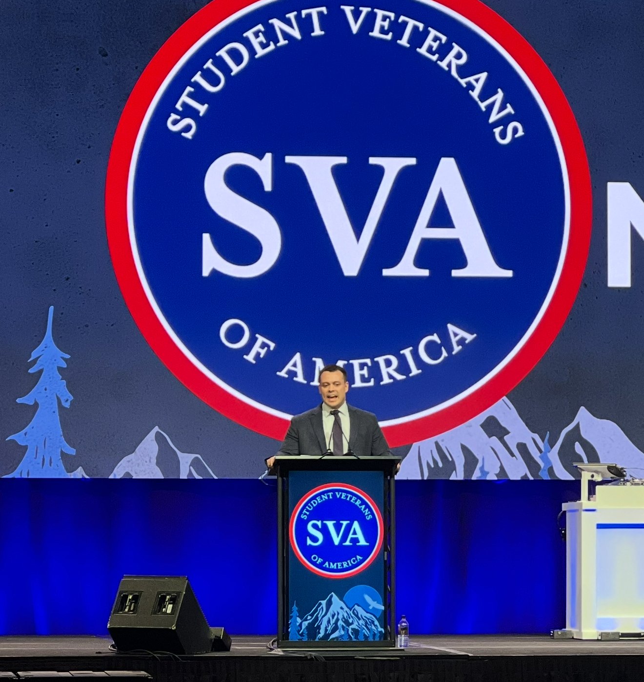 A man in a dark suit and tie stands behind a podium giving a speech at a conference with a large screen behind him displaying a logo that reads 'STUDENT VETERANS OF AMERICA' and the acronym 'SVA'.