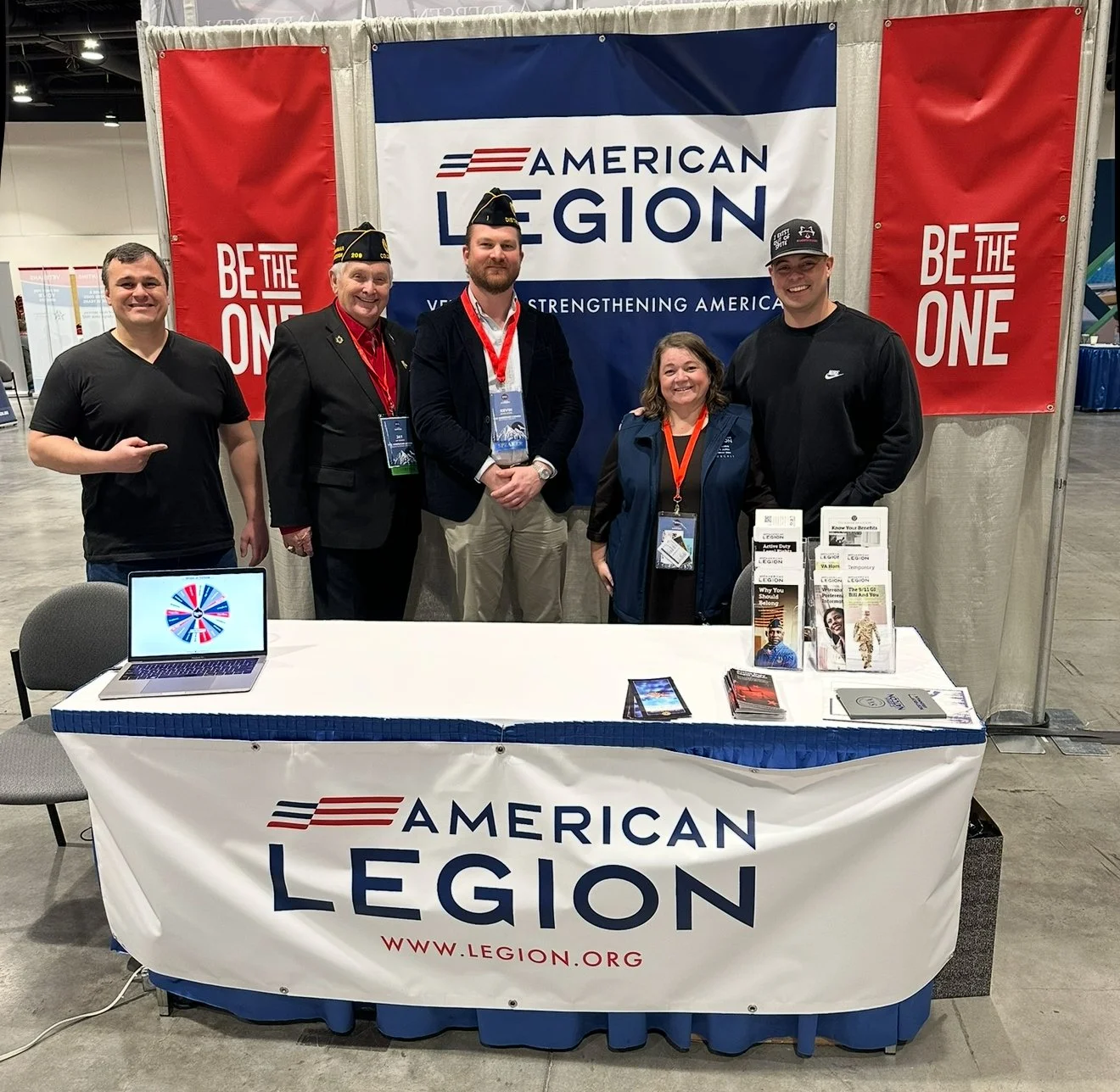 Group of five people standing behind an American Legion booth at an event, with a large American Legion banner and red side banners that read People the One. The booth has pamphlets, a tablet, and various flyers displayed on the table.