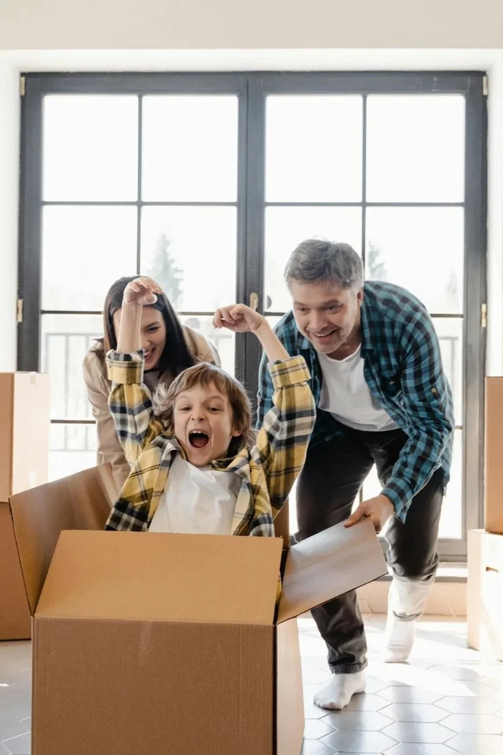 A family of three, a boy, a woman, and a man, playing and celebrating around a cardboard box in a room with large windows happy for their impact-resistant windows and doors in Tampa Bay Area single-family homes