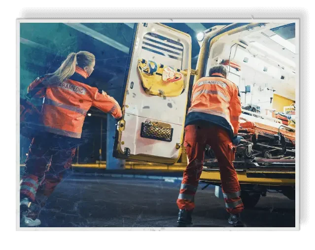 Two paramedics in orange uniforms loading a patient on a stretcher into an ambulance during night time showing the risk of injury without impact resistant windows and doors in businesses and homes in the Tampa Bay Area
