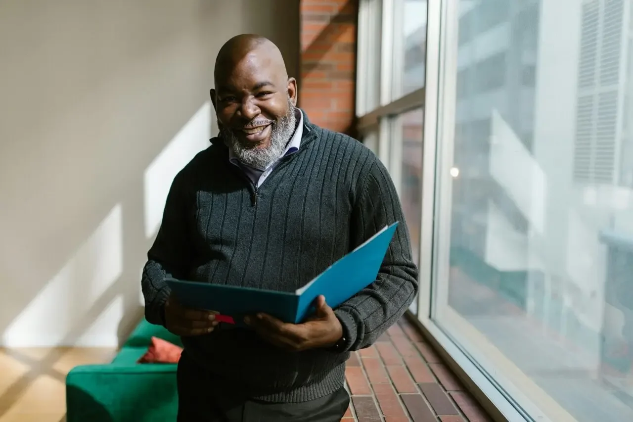 An older man with a beard, smiling and holding an open blue folder, standing near large windows in a historic building in the Tampa Bay Area  happy for their impact-resistant windows and doors.