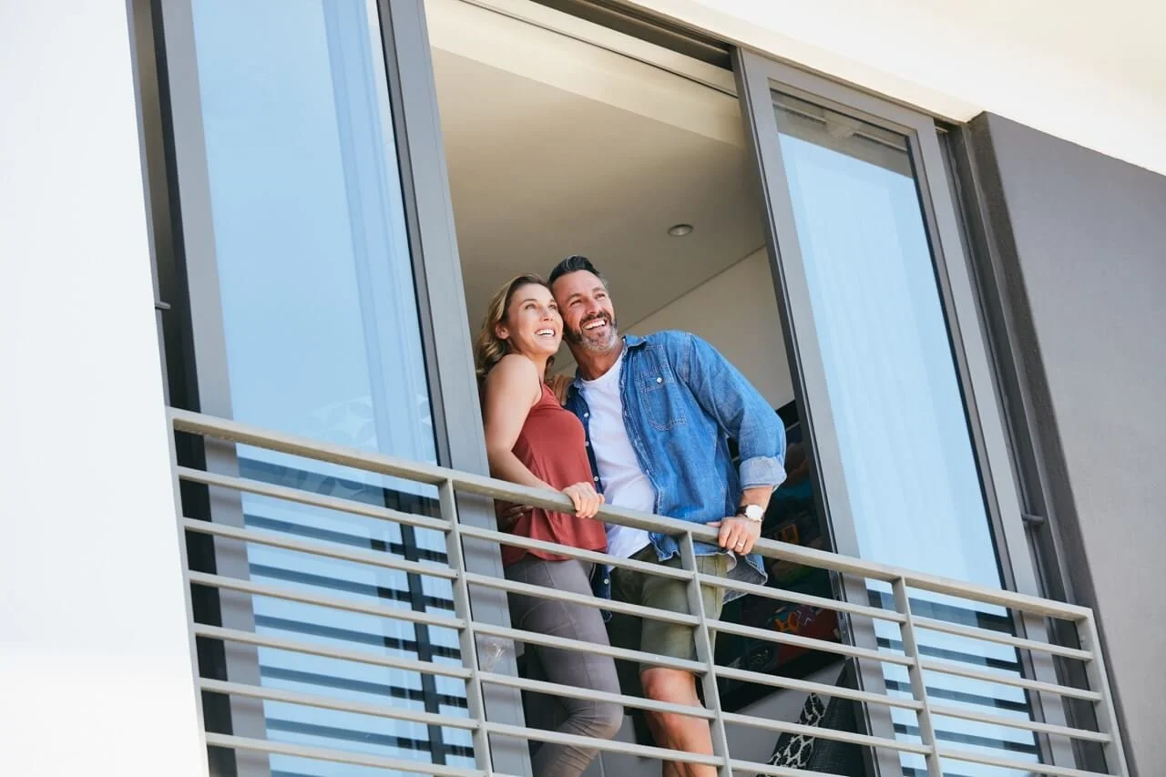 A smiling couple leaning out of a balcony door of their condo happy for their impact-resistant windows and doors in the Tampa Bay Area