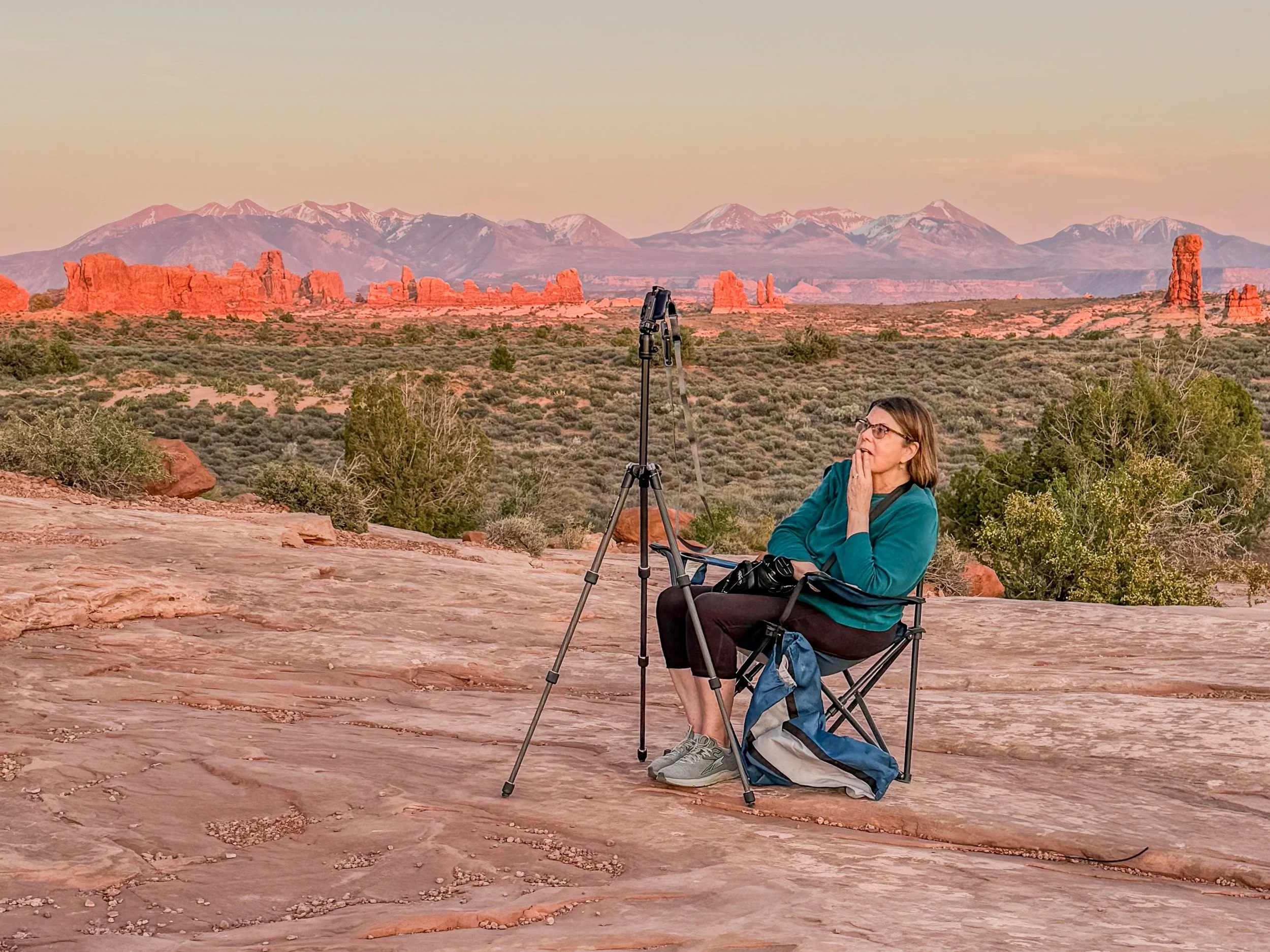A woman sitting on a camping chair outdoors, with a camera on tripod in front of her, overlooking a desert landscape with red rock formations, green shrubs, and snow-capped mountains in the distance during sunset.