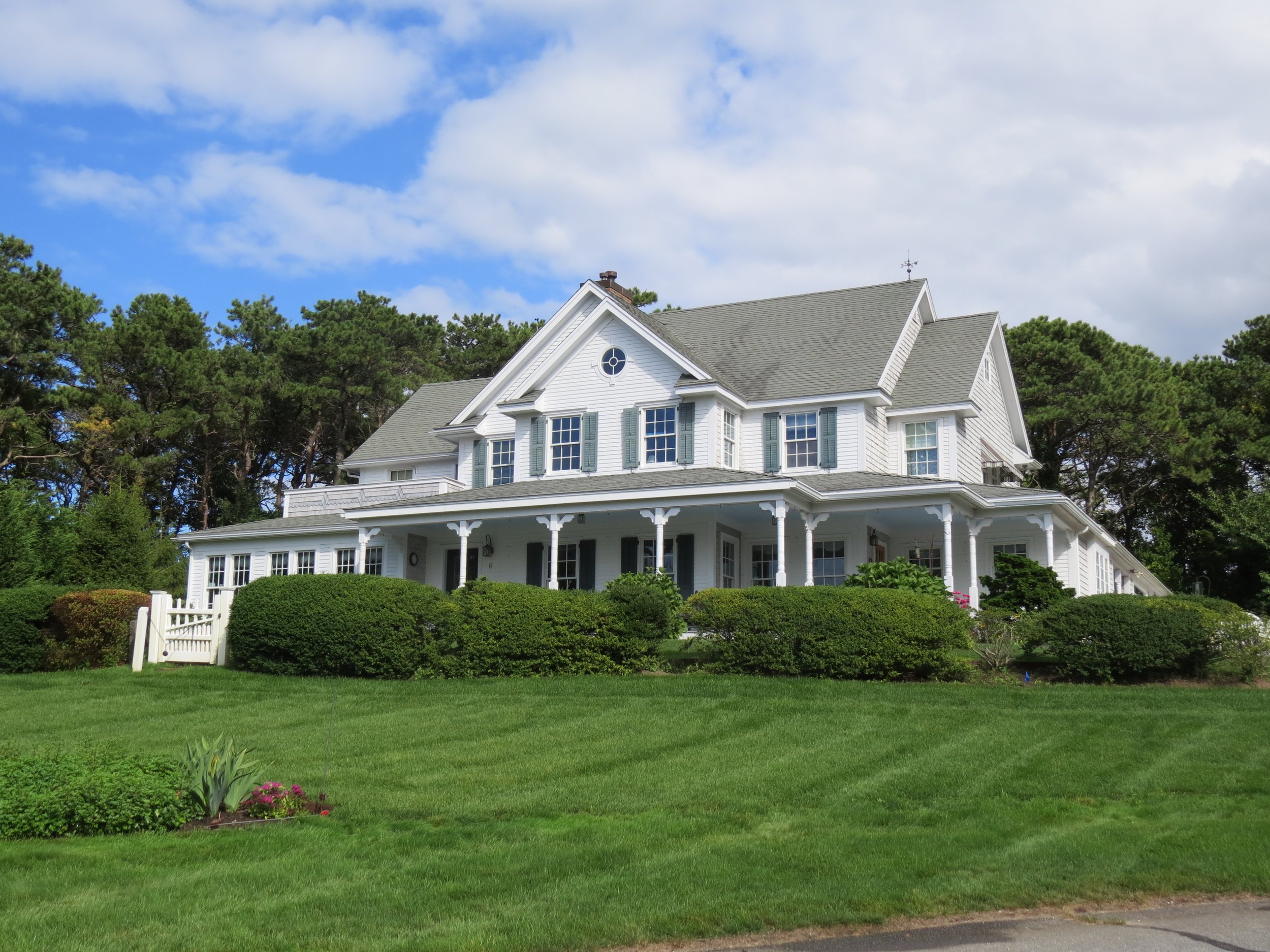 A large white house with multiple gabled roofs, surrounded by green bushes and a well-maintained lawn under a partly cloudy sky.