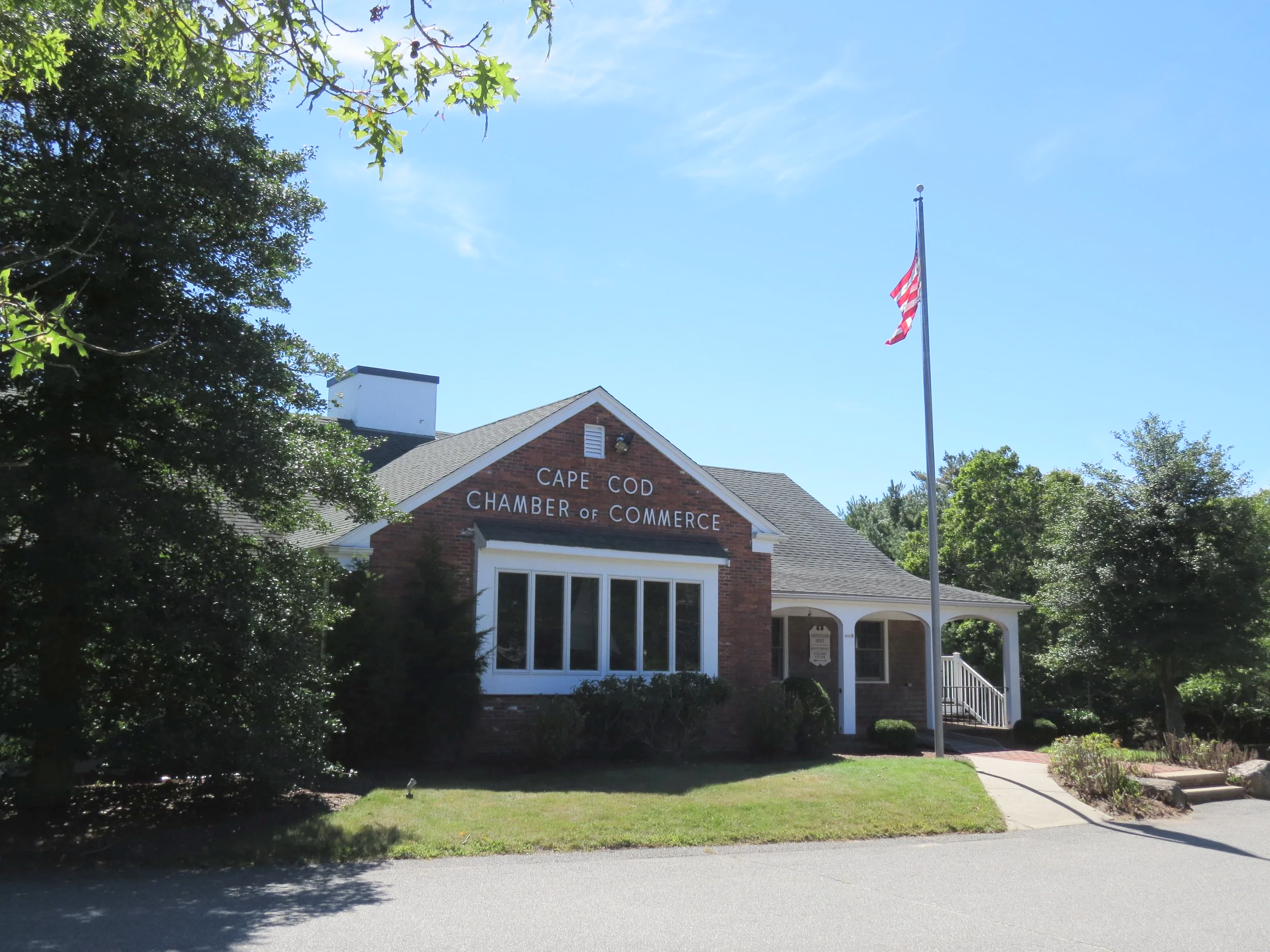 A brick building with a sign that reads 'Cape Cod Chamber of Commerce' and an American flag flying on a flagpole in front, surrounded by trees and a clear blue sky.