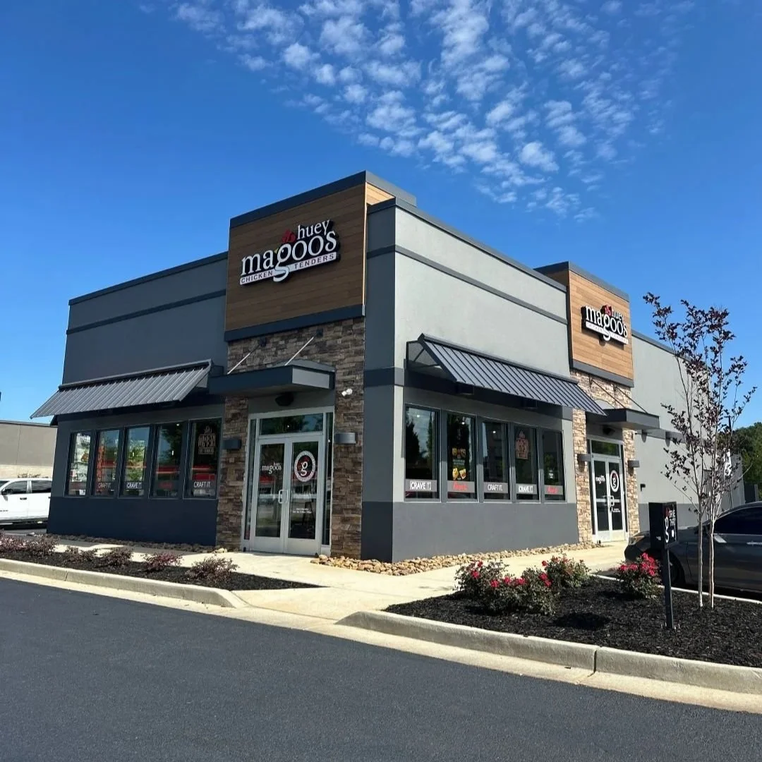 Exterior view of a modern fast-food restaurant named Huey Magoo's, specializing in chicken tenders, with a large sign, glass windows, and a parking lot with some cars.