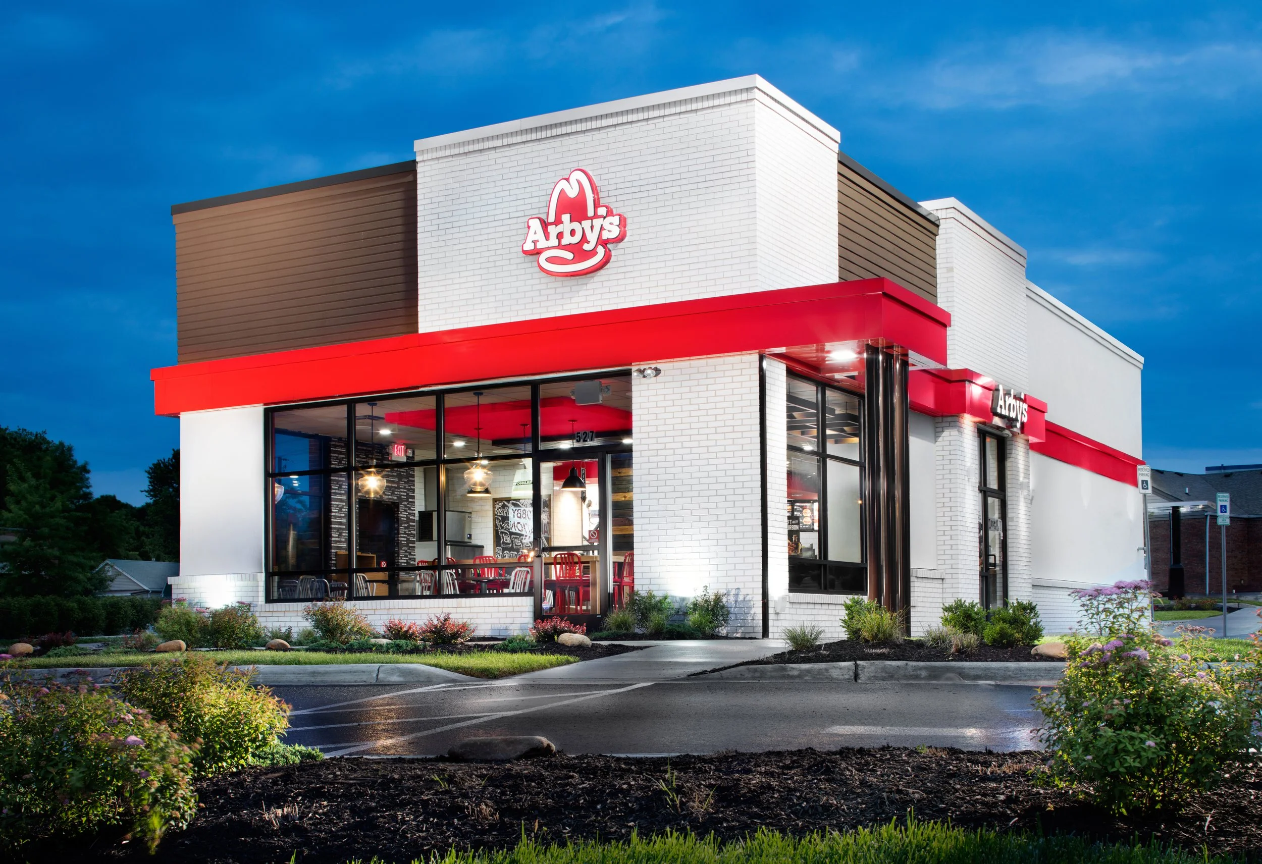 Exterior of a modern Arby's fast food restaurant at dusk, featuring large glass windows, white brick walls, and red signage, with a small landscaped parking lot in front.