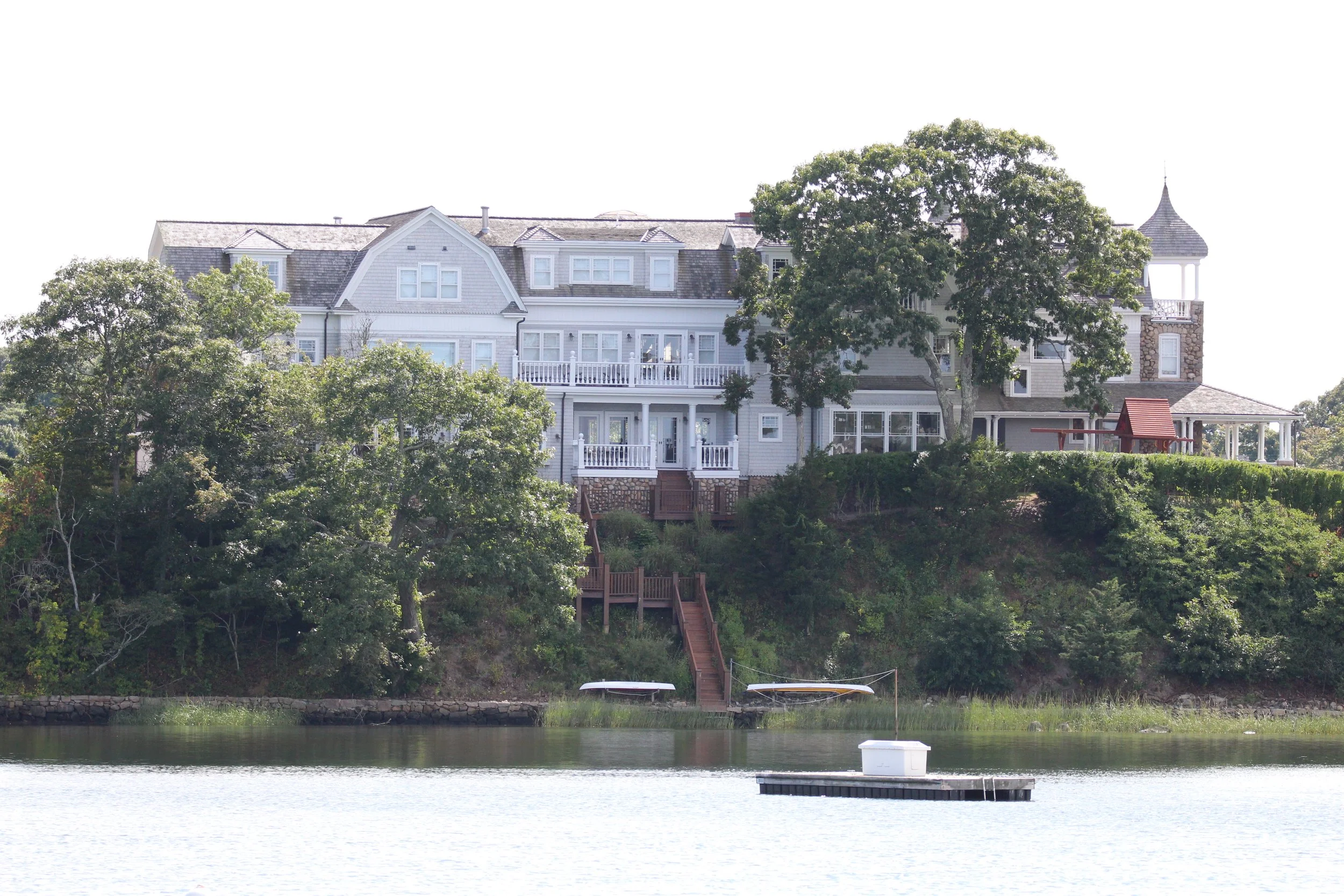 Large white house on a hill with multiple levels, surrounded by trees, overlooking a body of water with a dock and canoes.