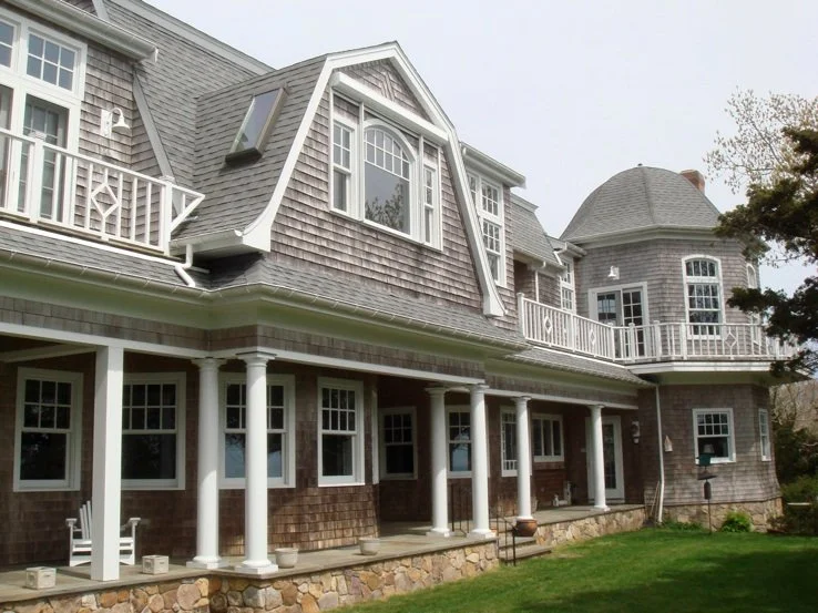 A large, multi-story house with gray shingle siding, white trim, and multiple windows, including a rounded turret and dormer windows, surrounded by green grass and some trees.