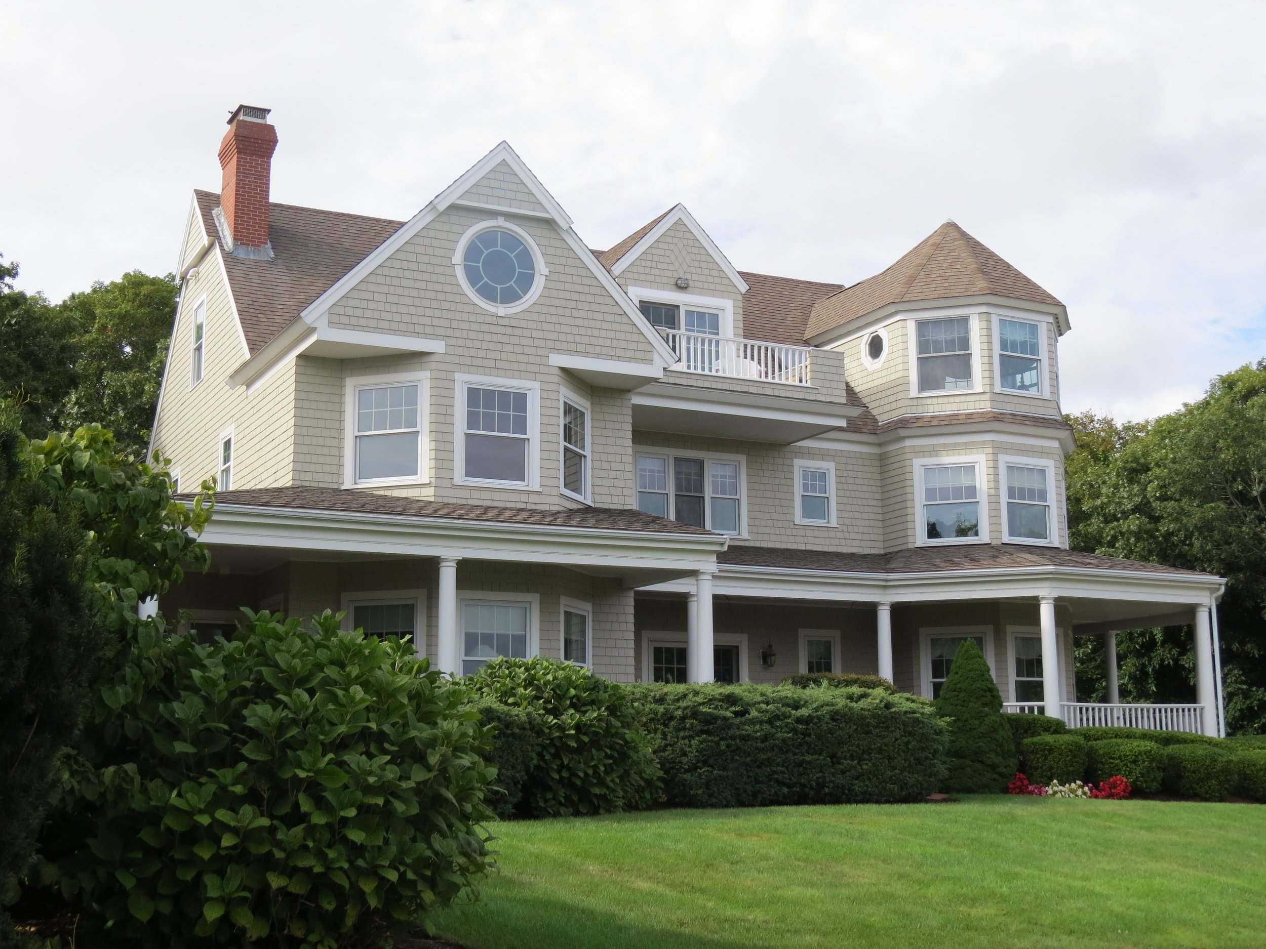 Large multi-story house with beige exterior, multiple windows, a porch, and a well-manicured lawn with bushes and flowers.