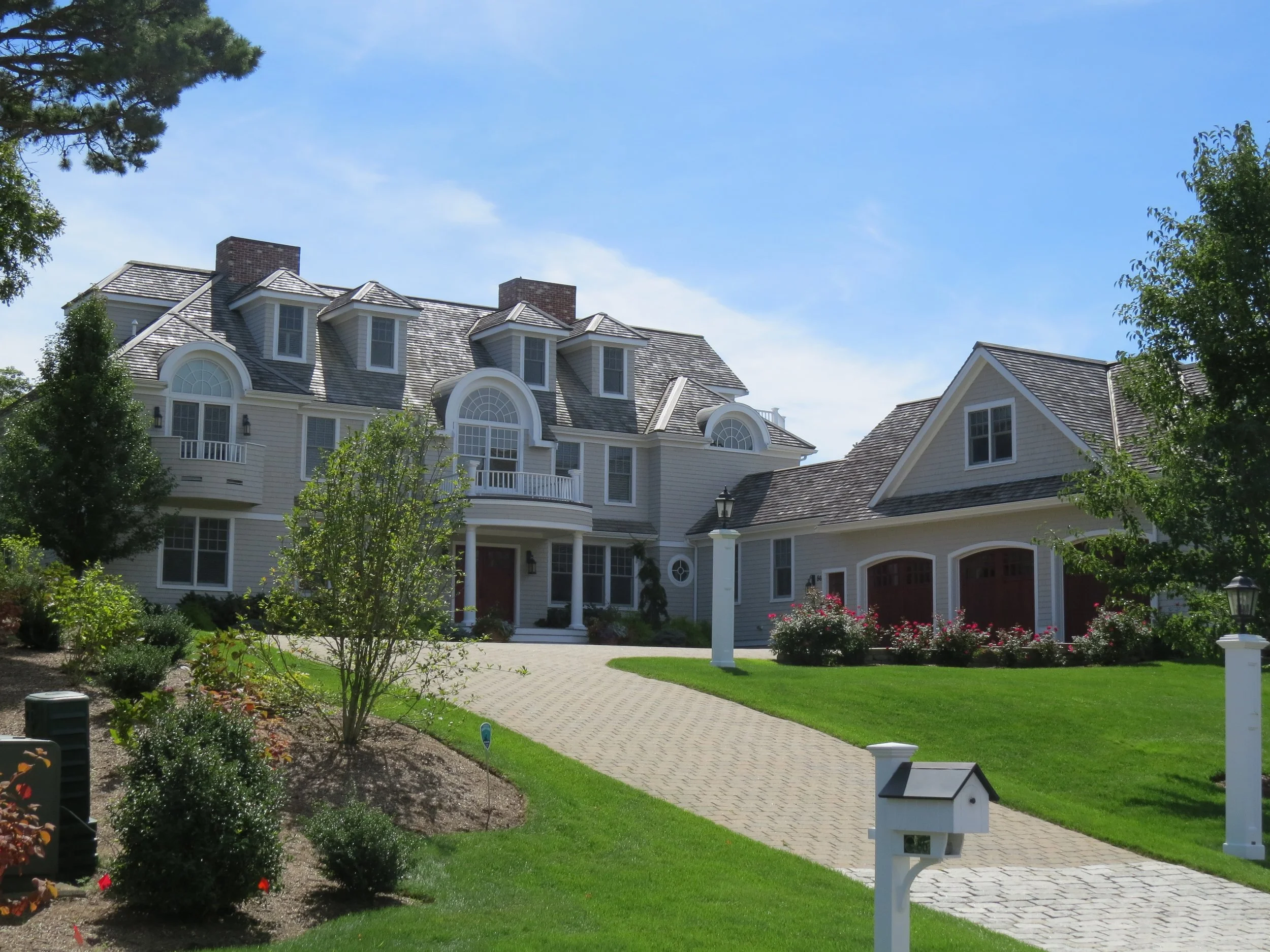 Large white multi-story house with dormer windows, surrounded by green lawn and landscaping, under a clear blue sky.