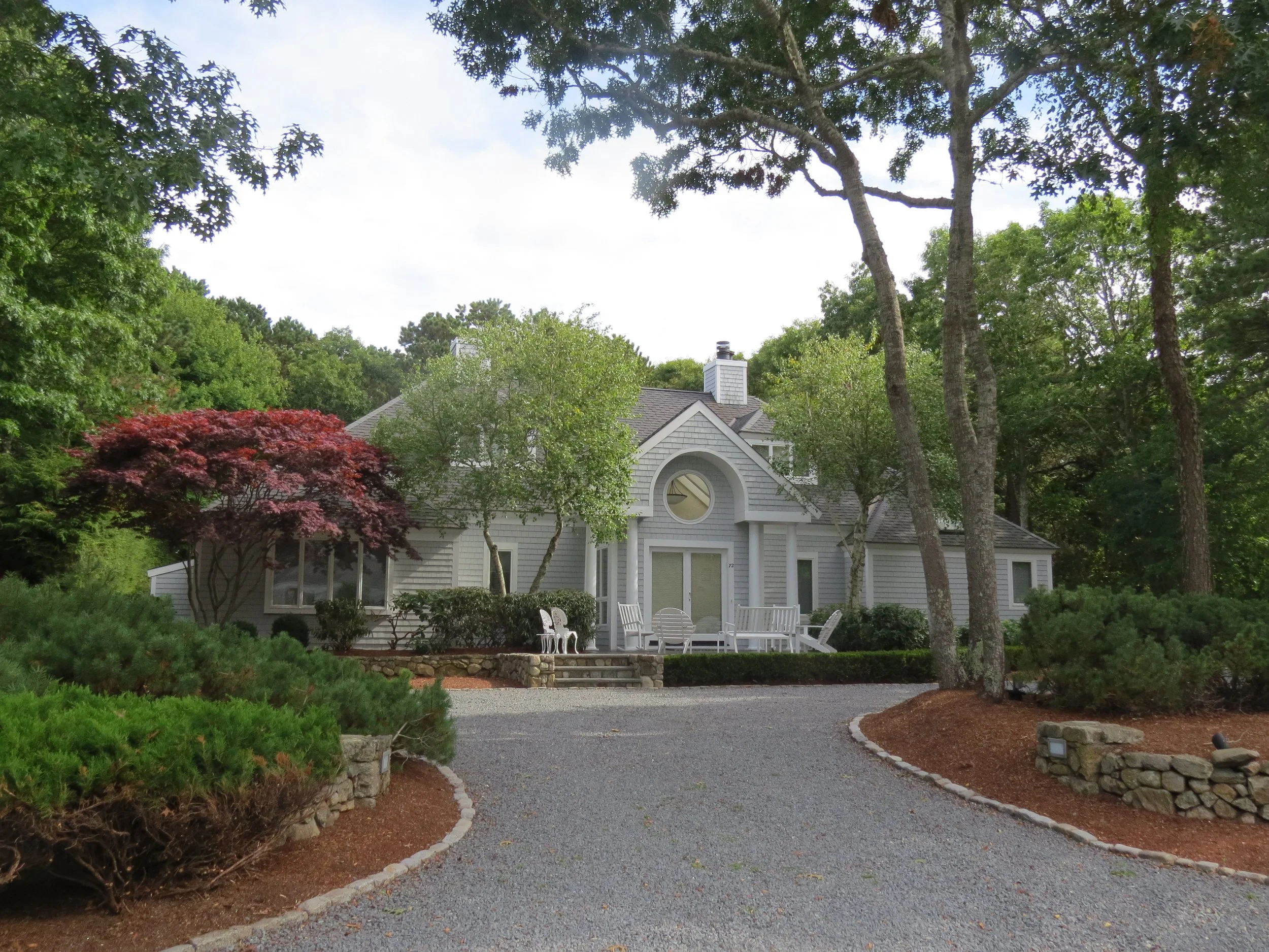 A house with gray siding and a gabled roof, surrounded by trees and shrubbery, with a gravel driveway leading up to it and outdoor white chairs on the front porch.