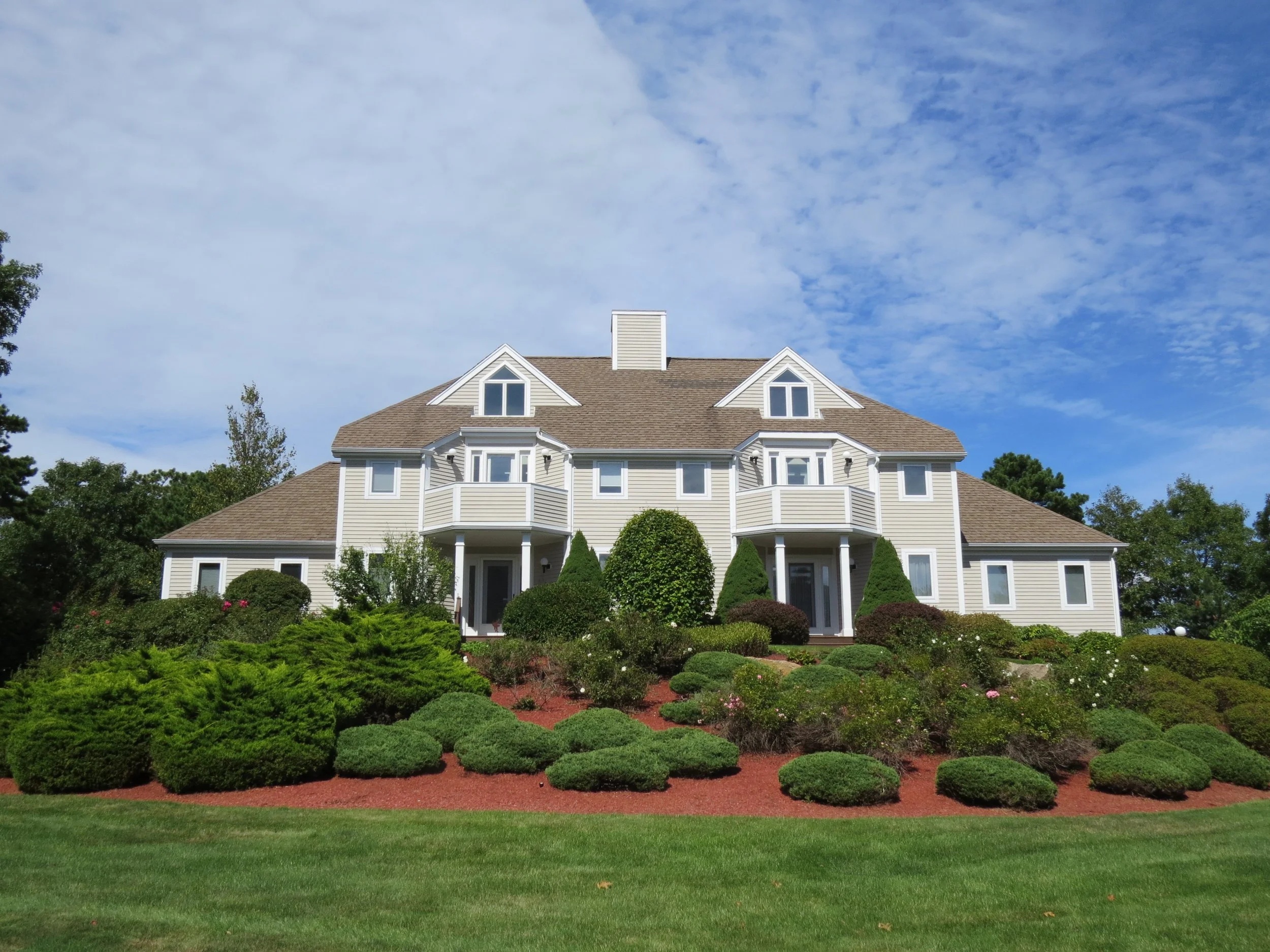 Large, white, multi-story house with a peaked roof and small balconies, surrounded by well-maintained garden with shrubs and trees, under a partly cloudy blue sky.