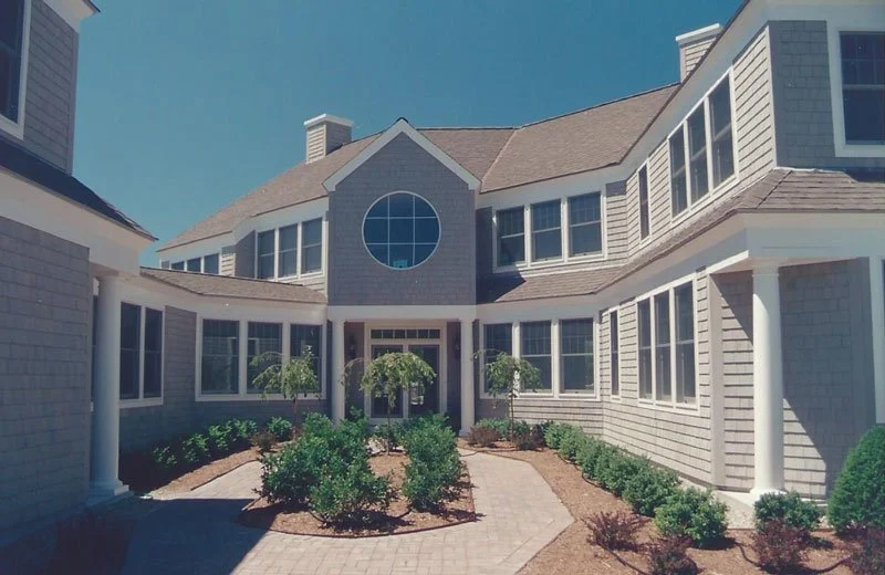 A large beige multi-story house with numerous windows and a circular window near the roof peak. A brick pathway leads to the entrance, flanked by small trees and bushes. The sky is clear and blue.