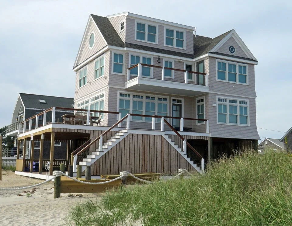A large, multi-story beach house with white siding, multiple windows, and balconies, located on sandy terrain with grass in the foreground.