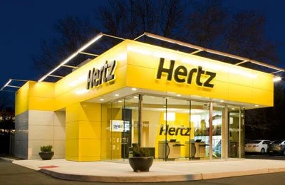 Hertz car rental store with a bright yellow exterior at dusk, large glass windows, and the Hertz logo displayed prominently.