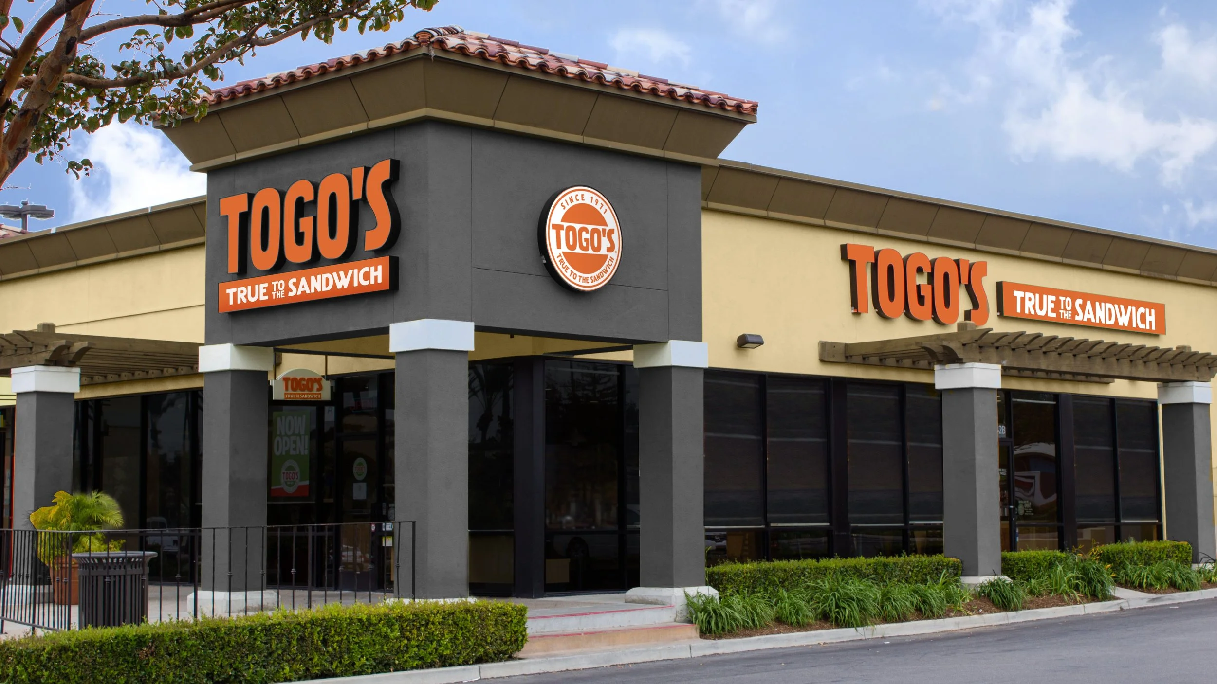 Exterior of a Togo's sandwich shop with large signs displaying the logo and slogan, with glass windows, a small patio with plants, and a clear sky.