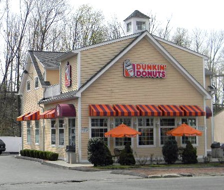 Dunkin' Donuts storefront with striped orange awning, outdoor seating with orange umbrellas, and the Dunkin' Donuts sign on a beige building.