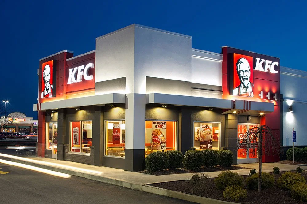 Exterior of a KFC restaurant during evening with illuminated signs, red, white, and gray building design, large windows, and a drive-thru