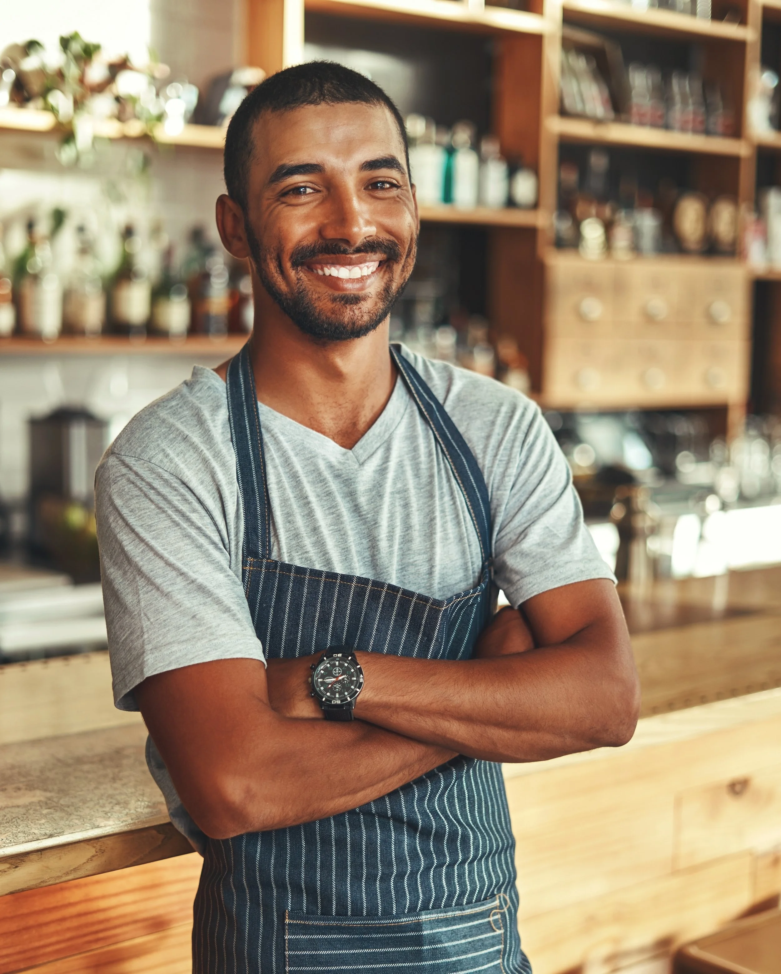 A smiling man with a beard and short hair standing behind a wooden counter in a cozy cafe or coffee shop, wearing a gray t-shirt and a blue striped apron.