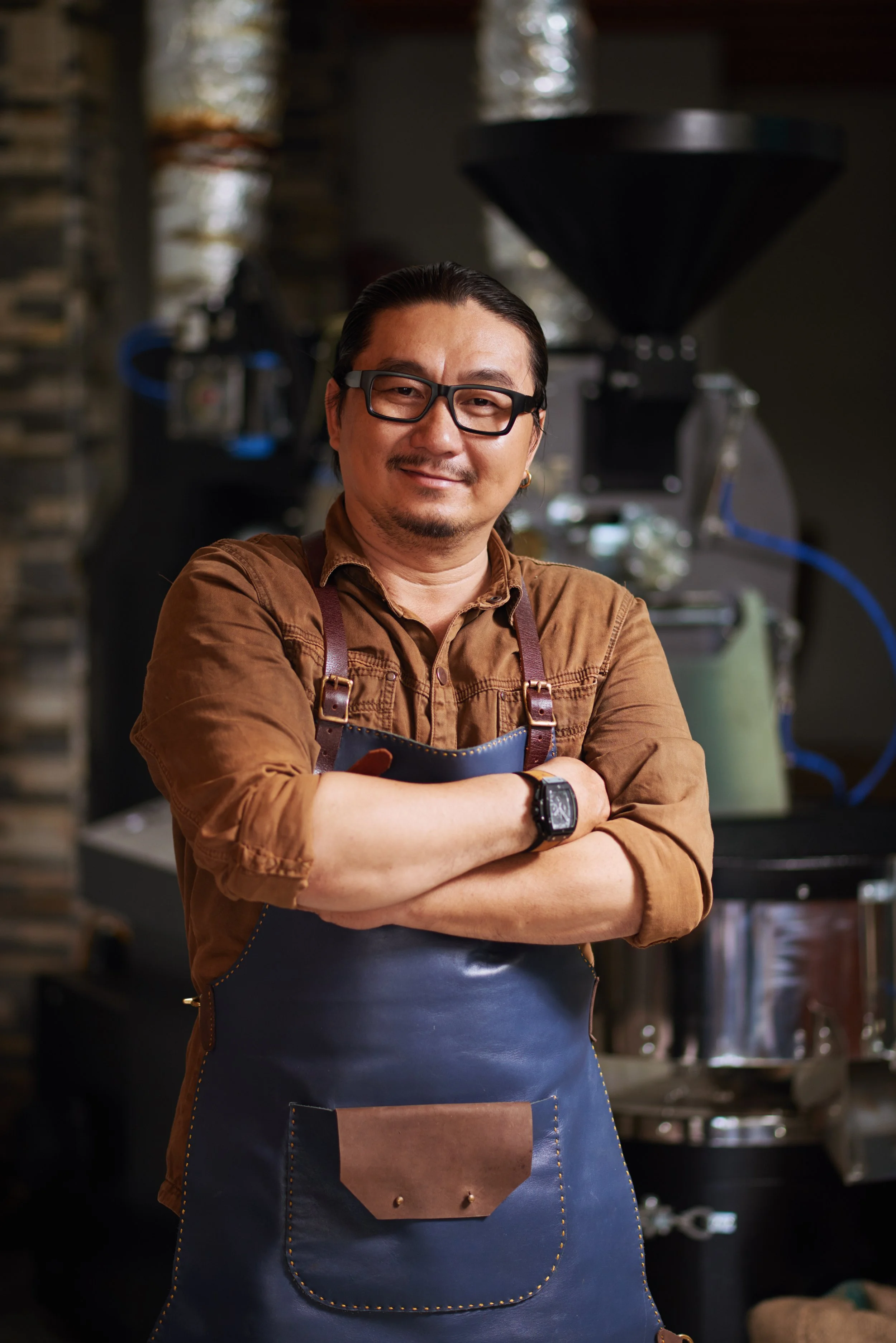 A smiling man with glasses and a mustache, wearing a brown shirt and a blue apron, stands with arms crossed in a workshop.