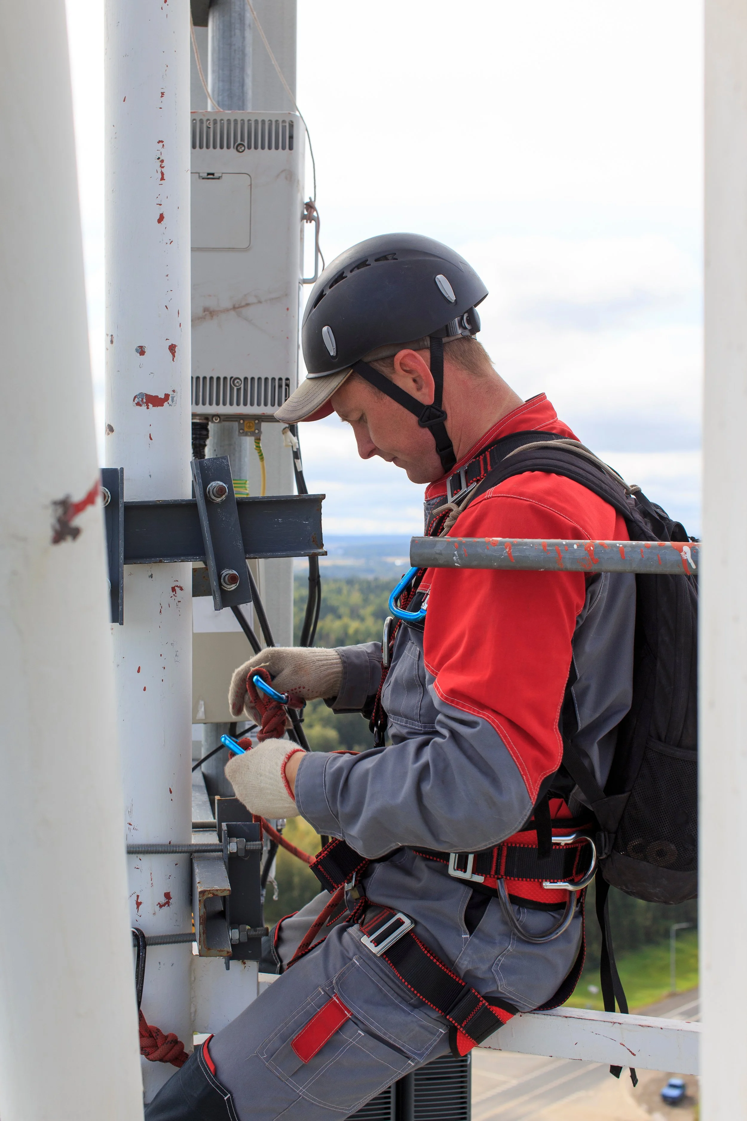 A male worker wearing a black safety helmet, gloves, and a backpack, working on electrical equipment on a utility pole outdoors.