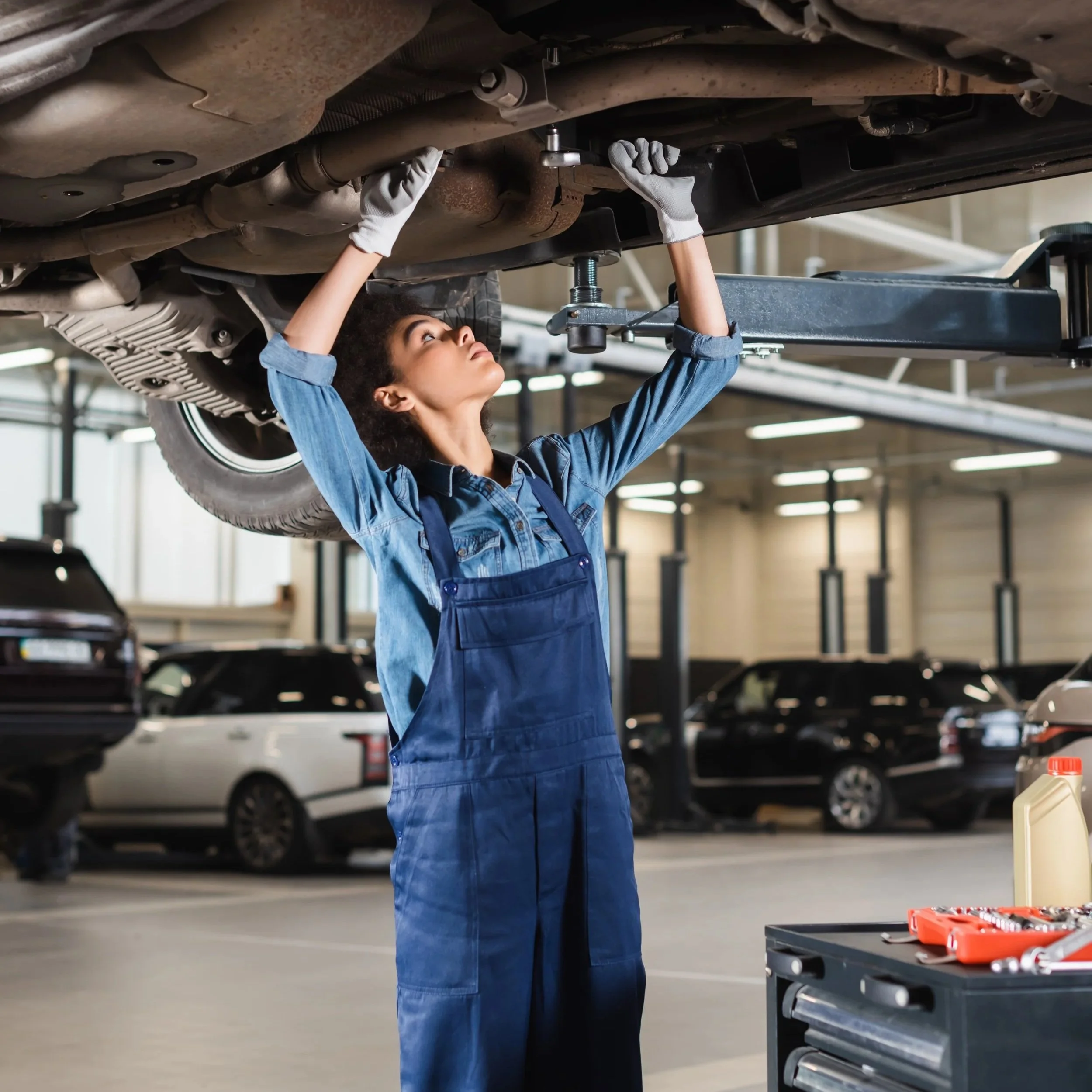 A woman working underneath a car in an automotive repair shop, wearing a blue jumpsuit and white gloves, with tools and a car lift visible.